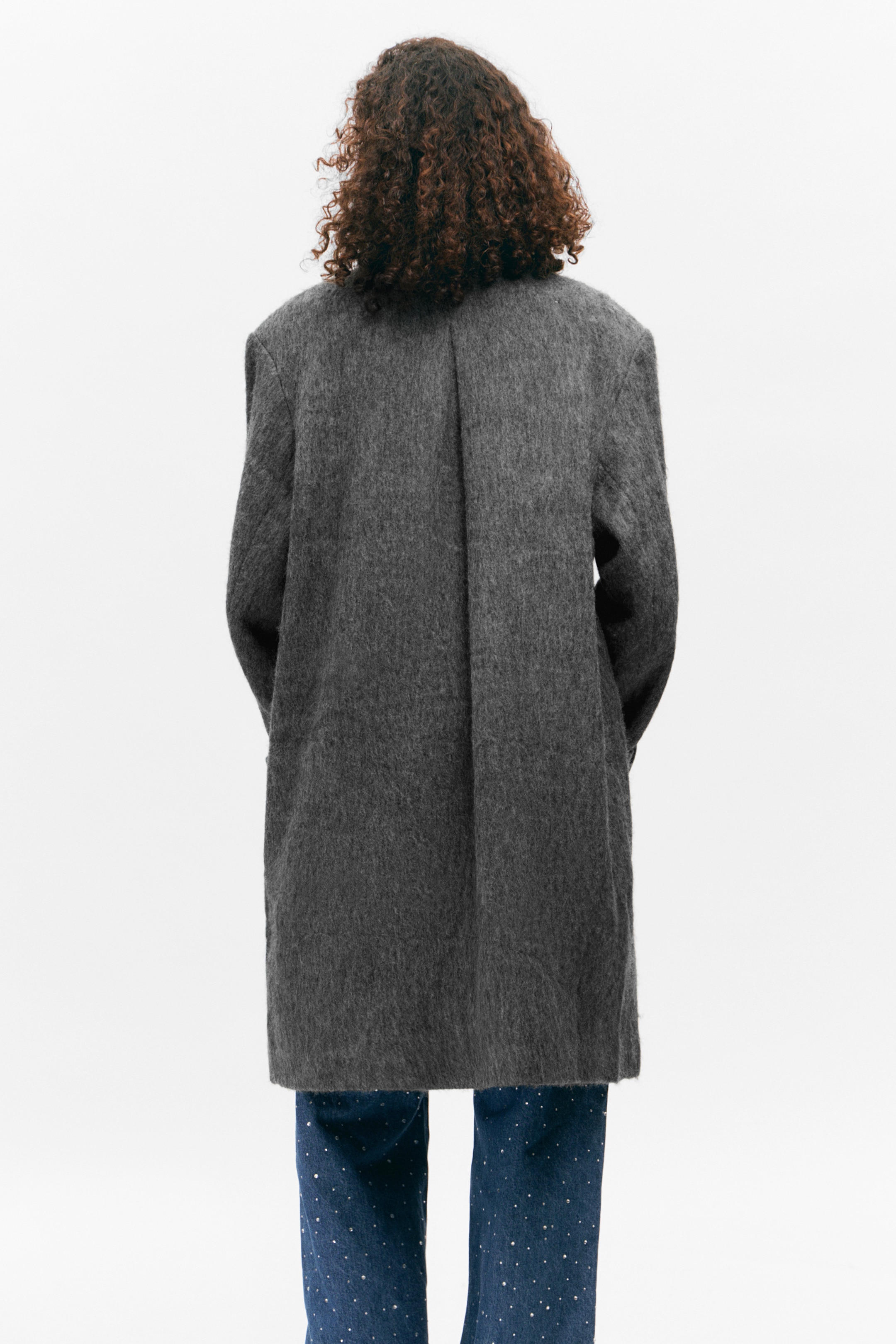 Agrandir l'image: Un manteau gris est présenté de dos, affichant une surface texturée et brossée avec une coupe décontractée. Il présente de longues manches et une couture centrale visible dans le dos, avec son ourlet s'étendant jusqu'à la partie supérieure de la cuisse.
