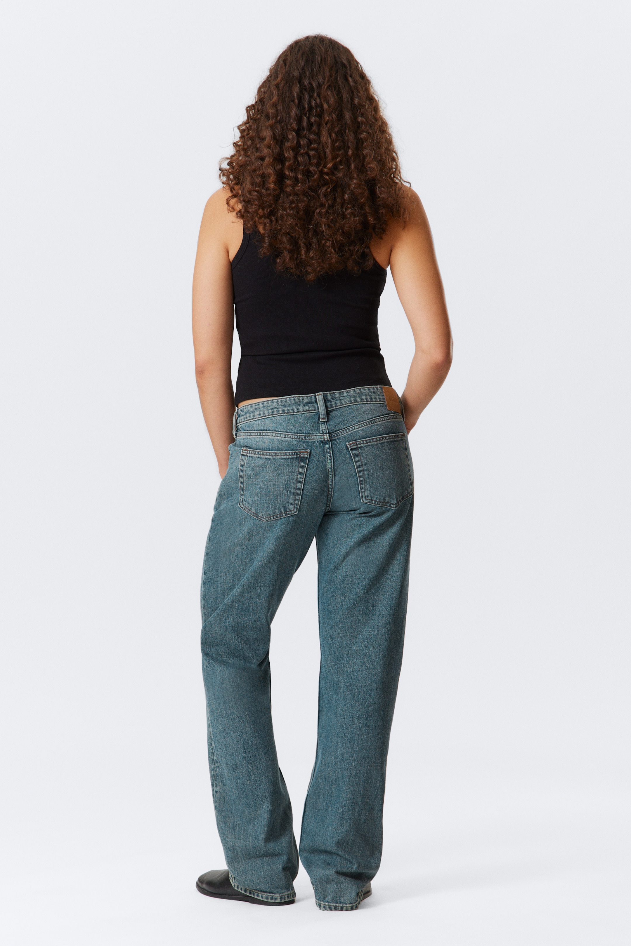 Grösseres Bild ansehen: Blaue Denim-Jeans werden von hinten präsentiert, mit einer lockeren Passform, zwei Gesäßtaschen und einem hellbraunen Aufnäher am Bund. Ein schwarzes geripptes Tanktop wird mit den Jeans getragen.