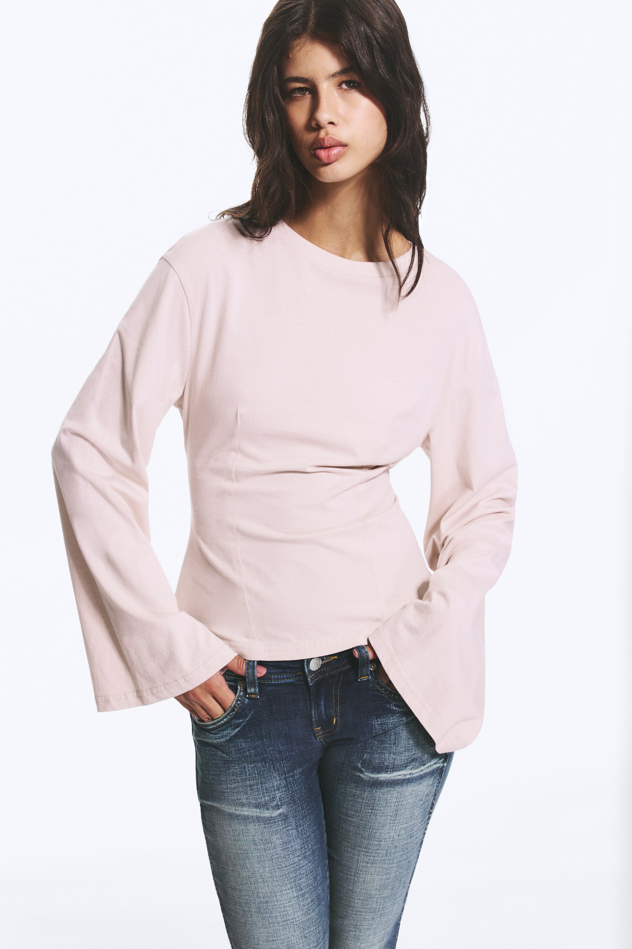 Visualizza immagine più grande: Una maglia a maniche lunghe di colore rosa chiaro presenta un girocollo e maniche ampie e svasate. È progettata con un punto vita aderente e cuciture verticali, creando una silhouette strutturata.