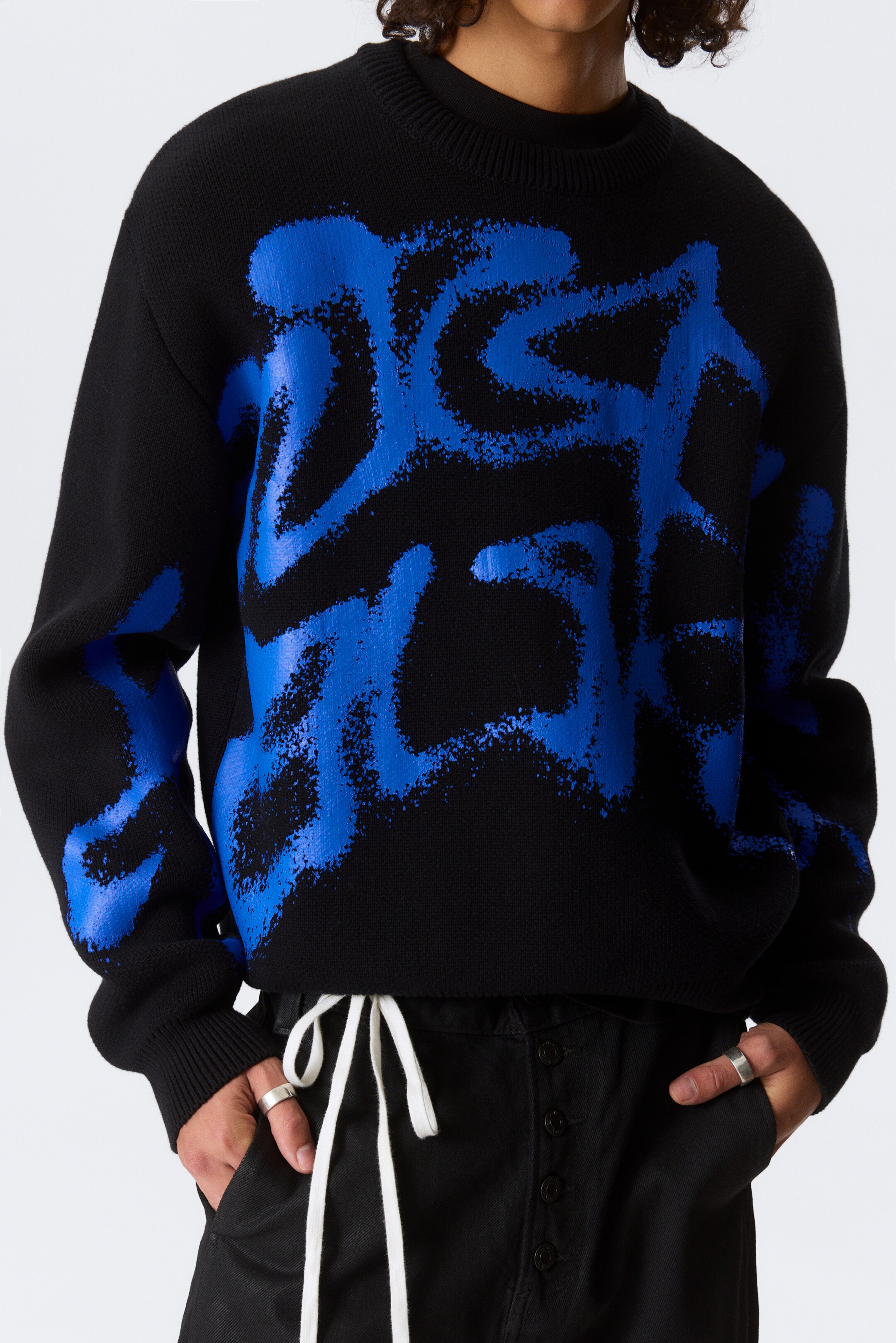 Se større billede: En sort sweater har en stor, abstrakt blå grafik, der ligner spraymaling, og som dækker forsiden og strækker sig ud på ærmerne. Sweateren har en ribbet rund hals, ribbede manchetter og en ribbet kant, og den bæres over en sort undershirt i en afslappet pasform, vinklet let til højre.