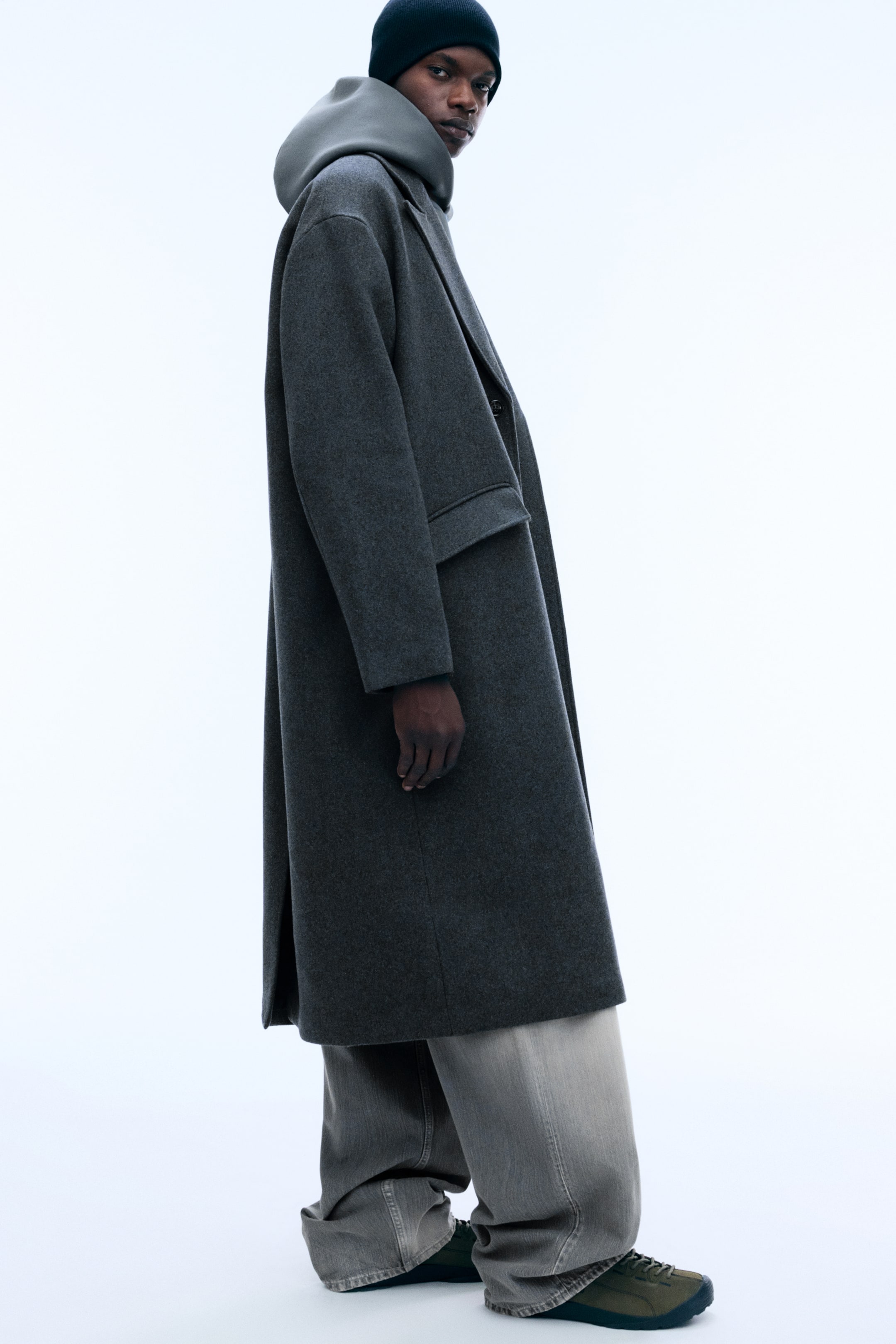 Ver imagem maior: Um casaco comprido em mistura de lã, de tom cinzento-escuro, com silhueta oversized e caída estruturada, é usado pela modelo numa vista de perfil. O casaco é sobreposto a uma sweatshirt cinzenta com capuz e jeans de perna larga, evidenciando o seu corte descontraído.