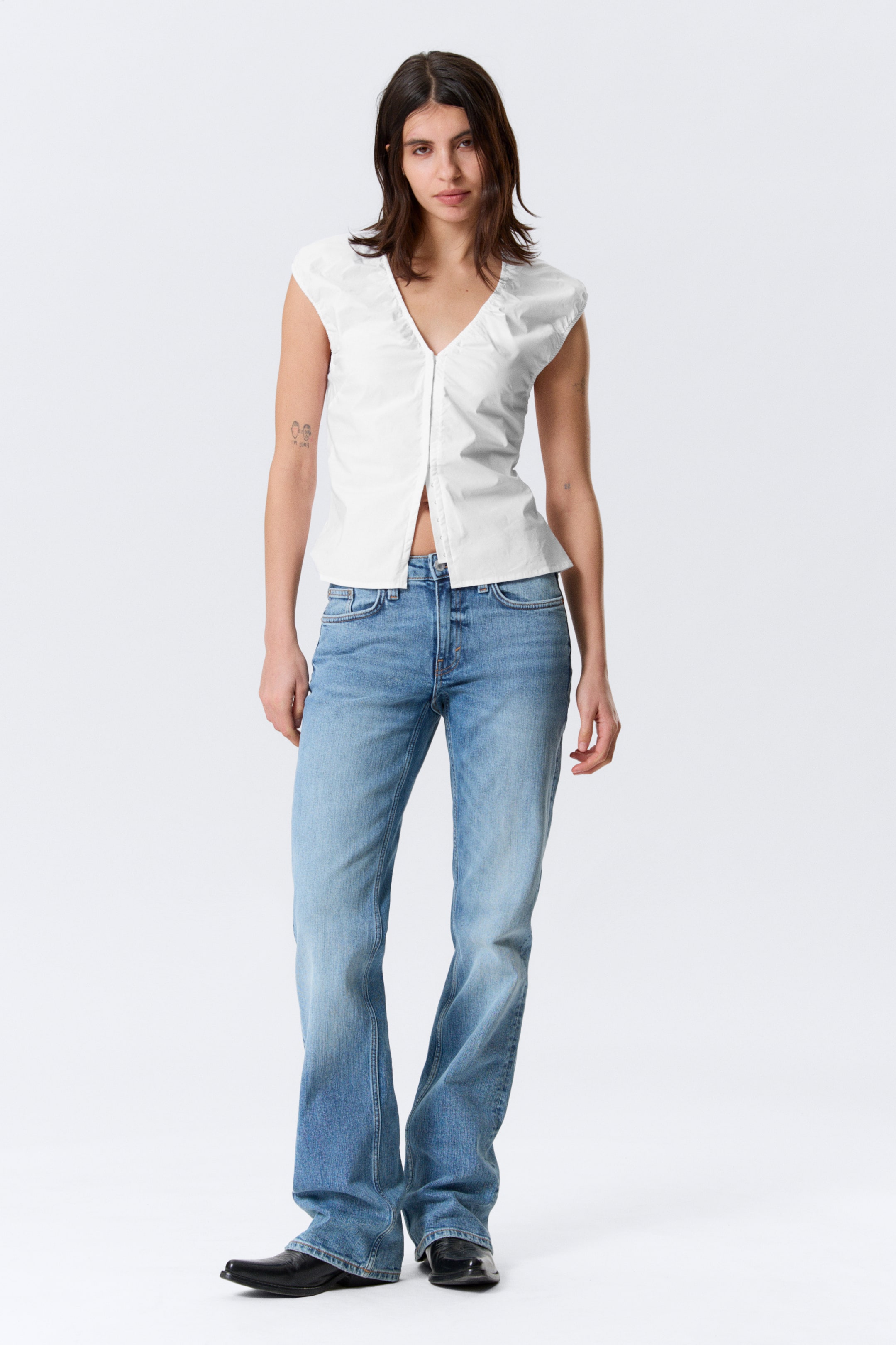 Ver imagen más grande: Un top blanco sin mangas con un escote en V, mangas abullonadas y cierre frontal de botones, dejando los botones inferiores desabrochados. Vaqueros de lavado azul claro con un ajuste de tiro medio y una ligera campana en la pierna.