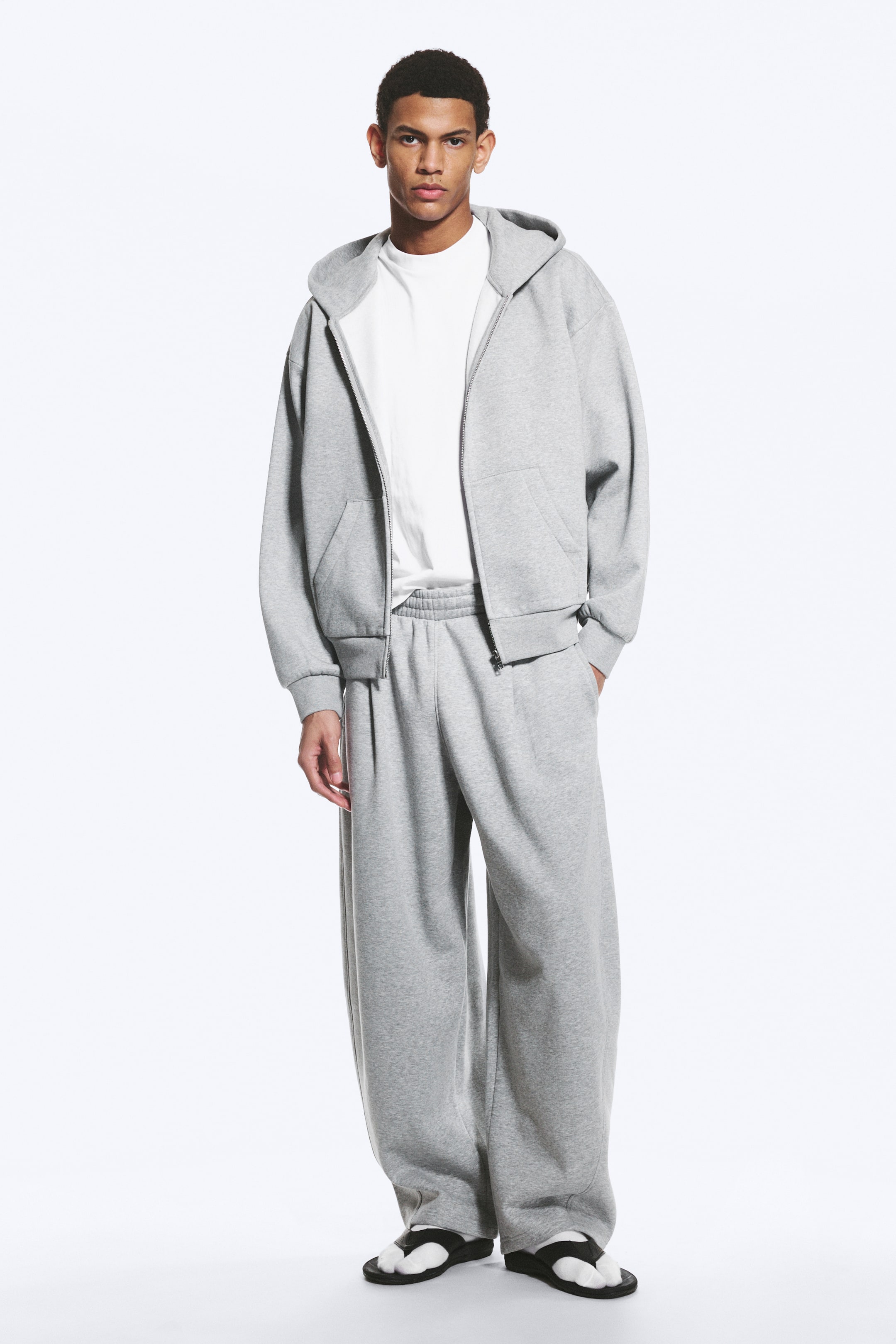 Ingrandisci l'immagine: Una felpa con cappuccio grigia con zip integrale, caratterizzata da un cappuccio, una cerniera frontale, tasche anteriori e orlo a costine, indossata sopra una t-shirt bianca. Sono visibili anche i pantaloni della tuta grigi a vestibilità ampia con un elastico in vita, presentando il completo da una prospettiva frontale.