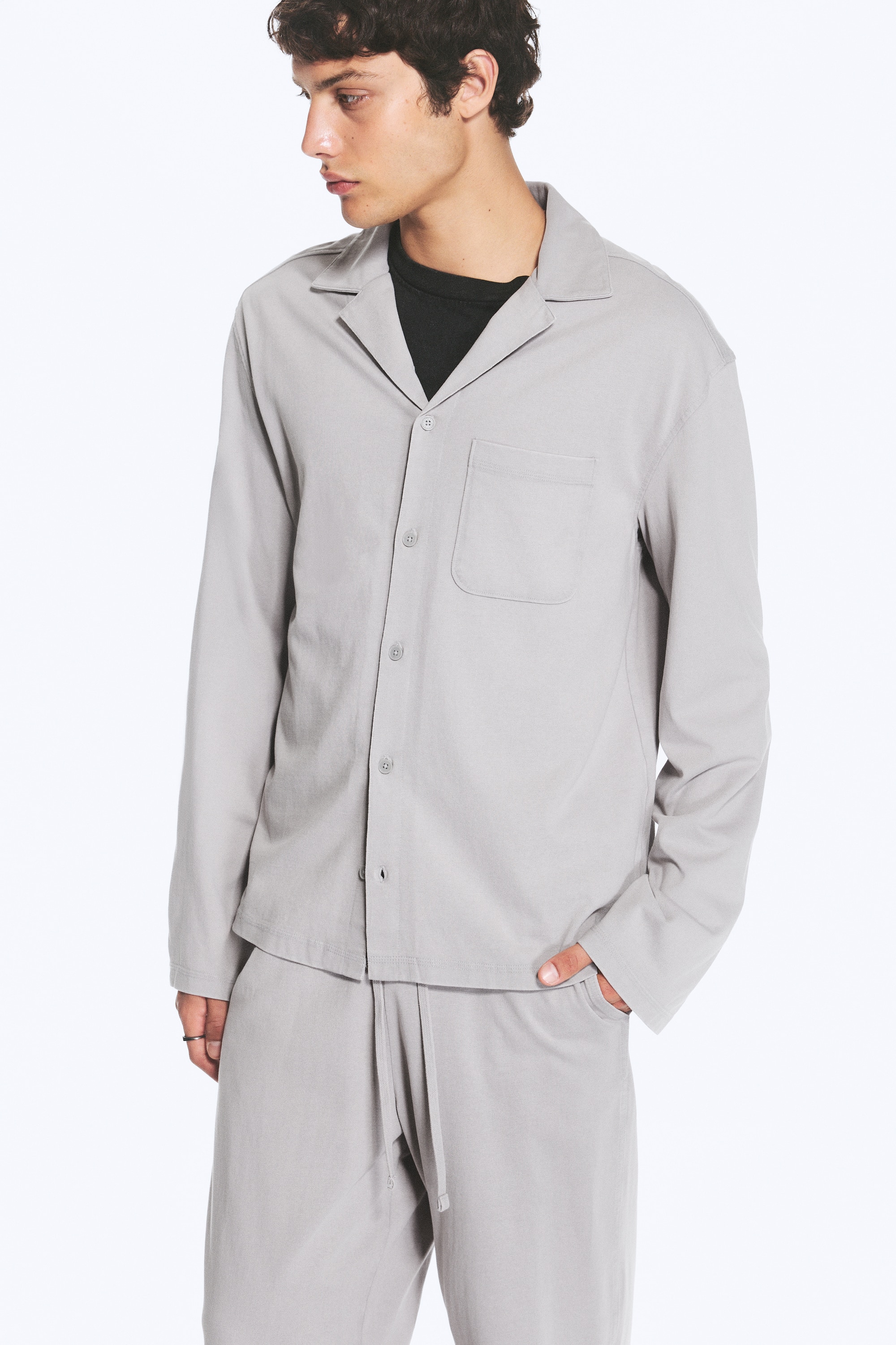 La camicia grigio chiaro, mostrata dal davanti, presenta un colletto a rever, una chiusura frontale con bottoni, maniche lunghe e rilassate, e una tasca applicata singola sul petto sinistro.