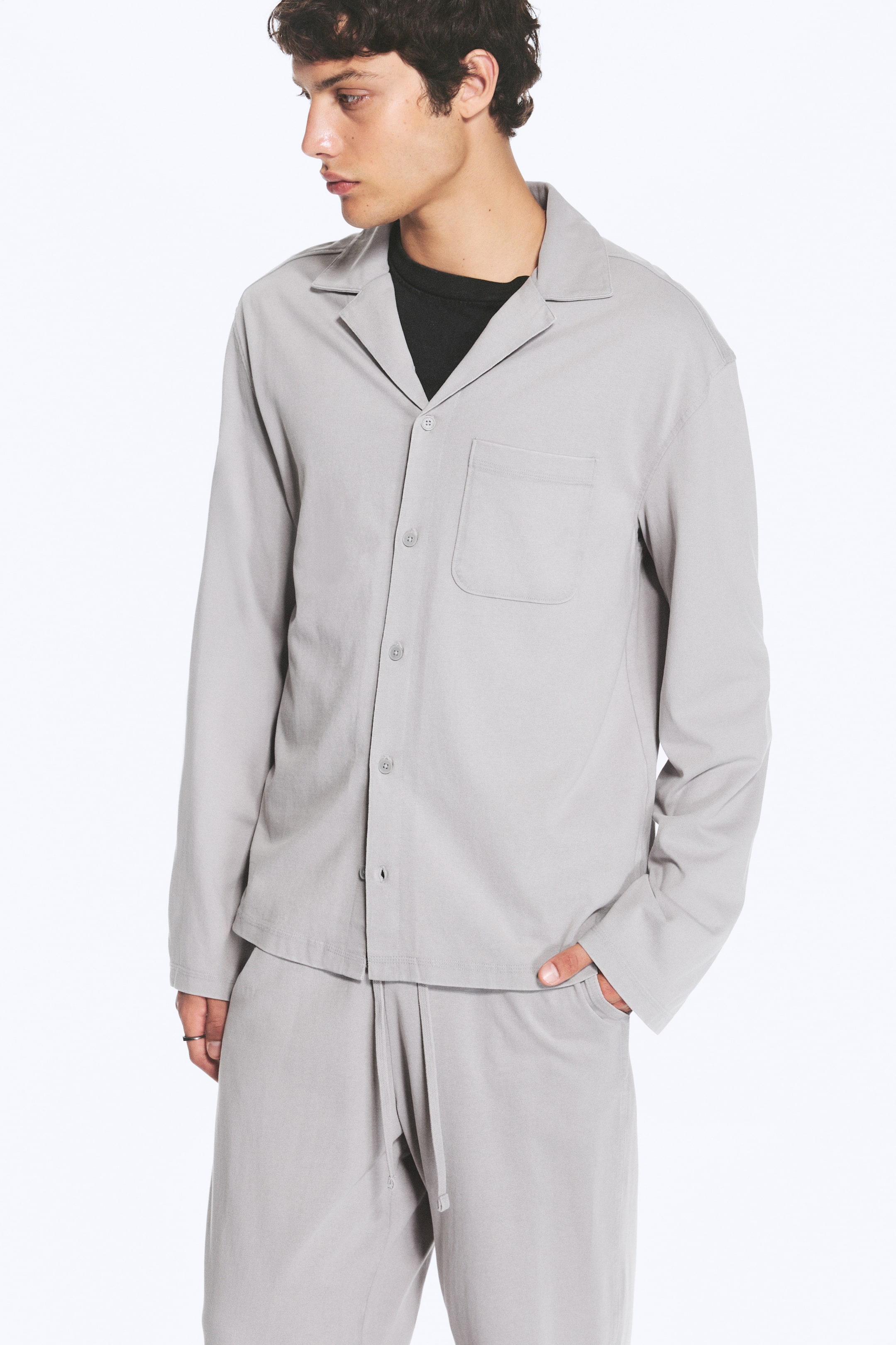 Vis større bilde: En lysegrå loungewear-skjorte med lange ermer, avslappet åpen krage, knepping foran og én brystlomme bæres av modellen. Skjorten er lagdelt over en svart T-skjorte og kombinert med matchende lysegrå bukser med snøring i livet.