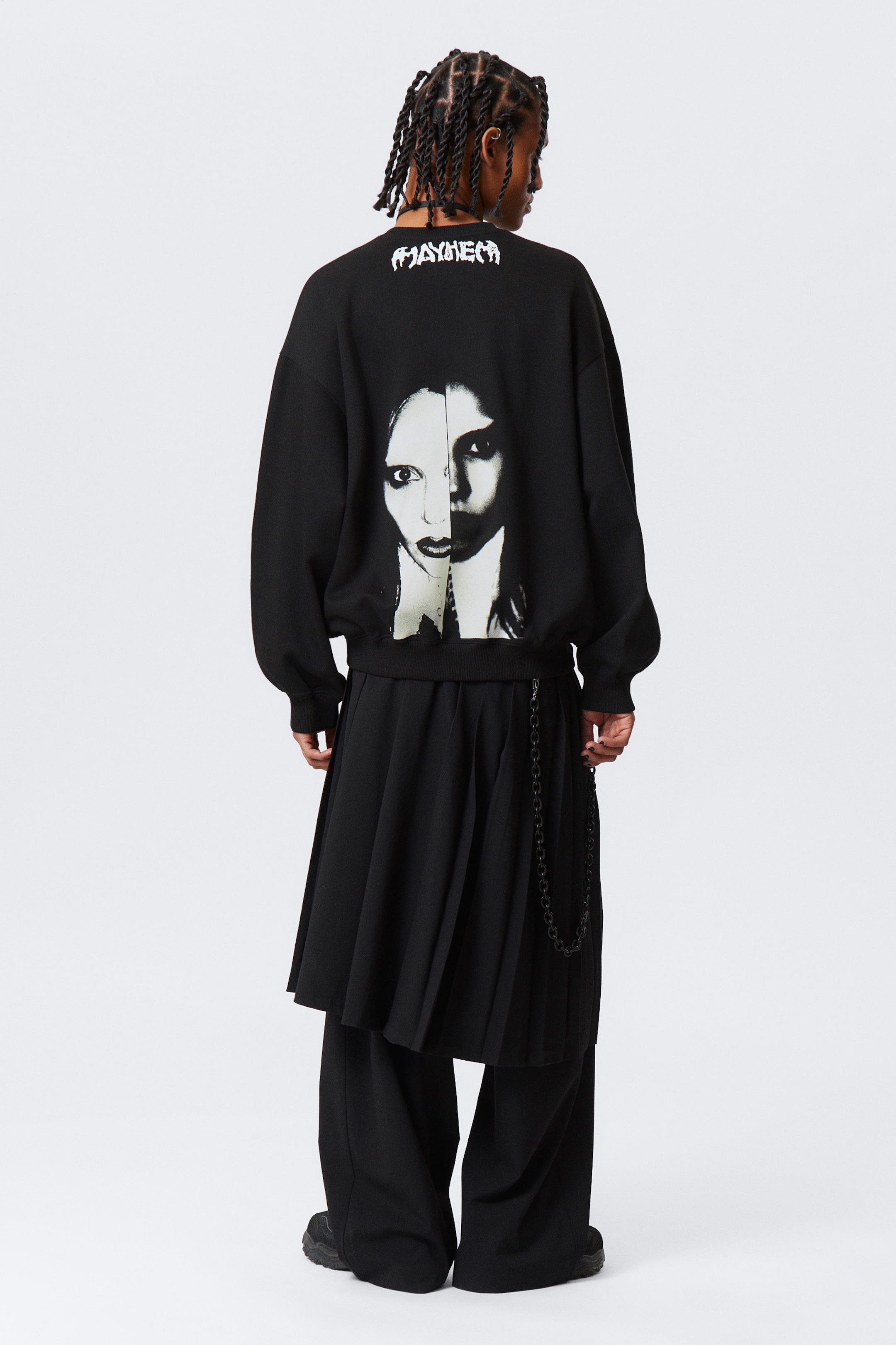 Agrandir l'image: Un sweat-shirt noir surdimensionné est présenté de dos, affichant une coupe décontractée avec des manches longues et un ourlet côtelé. Il présente un imprimé texte blanc "MAYHEM" sur le haut du dos et un grand graphique noir et blanc d'un visage fendu au centre de son dos, stylisé sur une jupe plissée noire et un pantalon large.