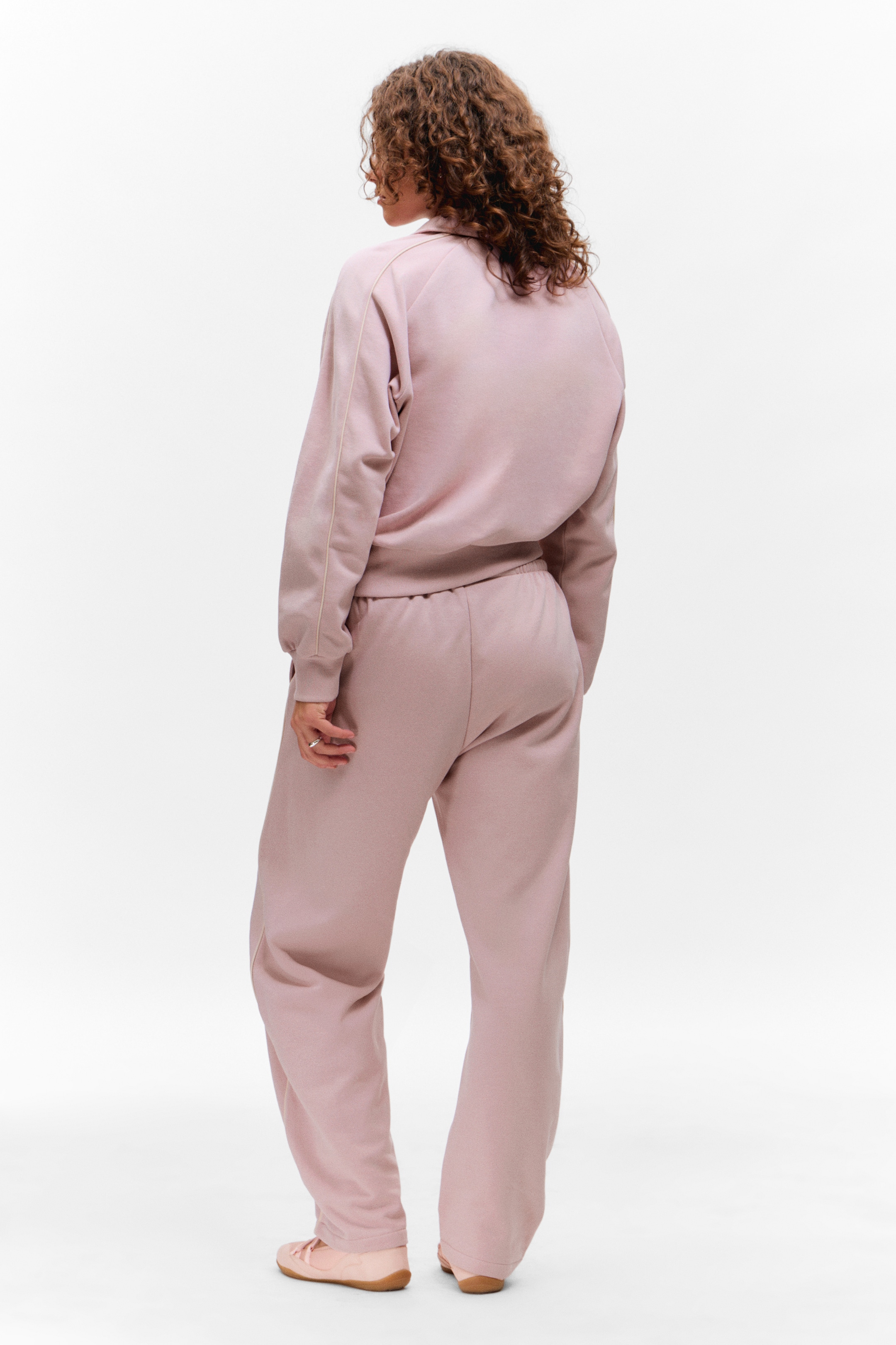 Vis større bilde: En dempet rosa joggedress blir sett fra en bakre, litt vinklet perspektiv. Toppen har en krage, raglanermer og en innsnevret kant, mens de matchende joggebuksene er i en vid stil med en elastisk linning.