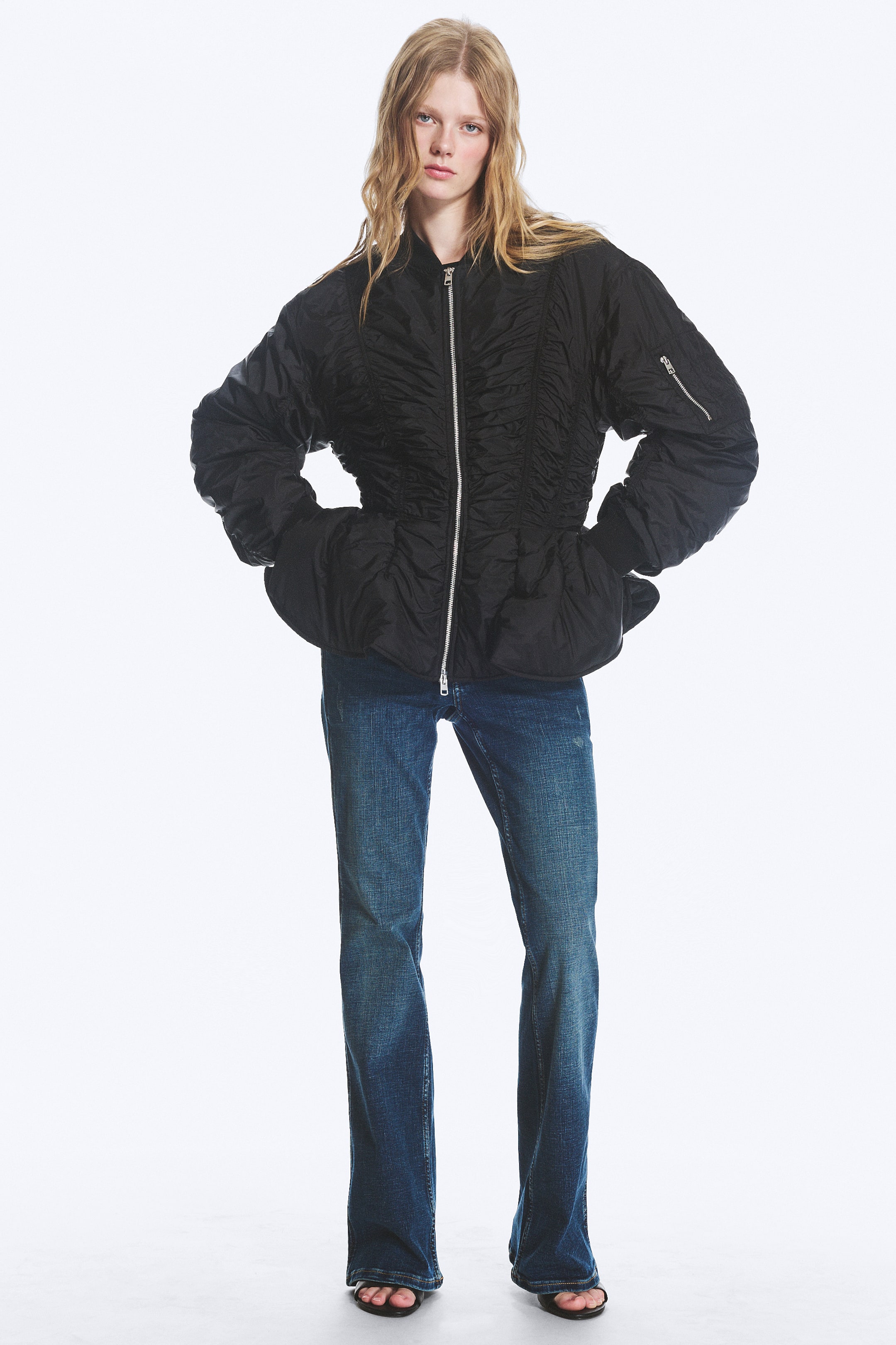 View larger image: A black bomber jacket featuring a two-way front zip, a zipped sleeve pocket, and all-over ruched seam detailing. The jacket boasts a peplum hem, with the model posing with her hands placed on her hips.