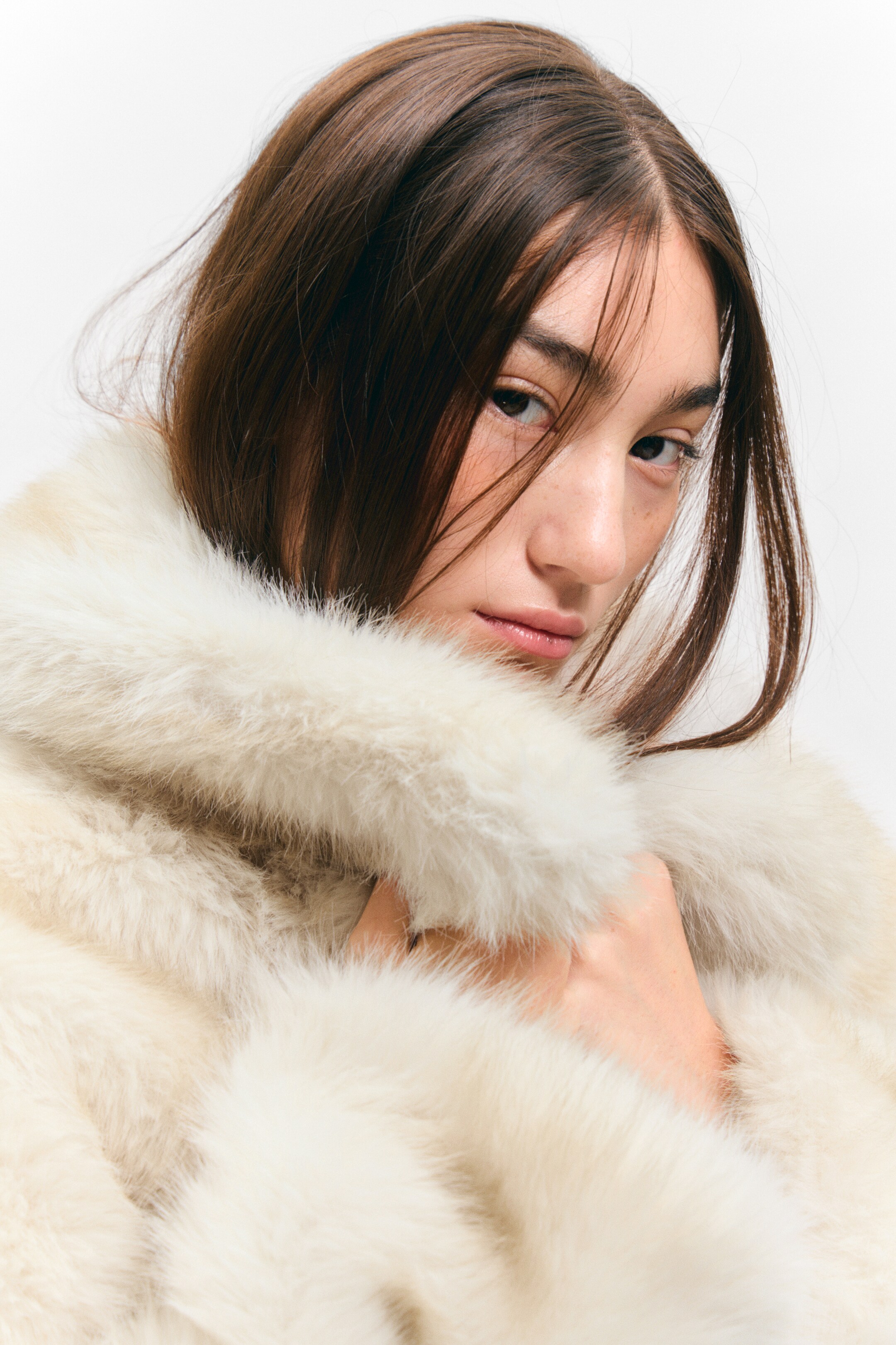 View larger image: Cream faux fur is framing a face in a close-up view, displaying a thick, plush texture around the collar and shoulder area. A hand is resting within the fur, showcasing its volume and soft appearance.