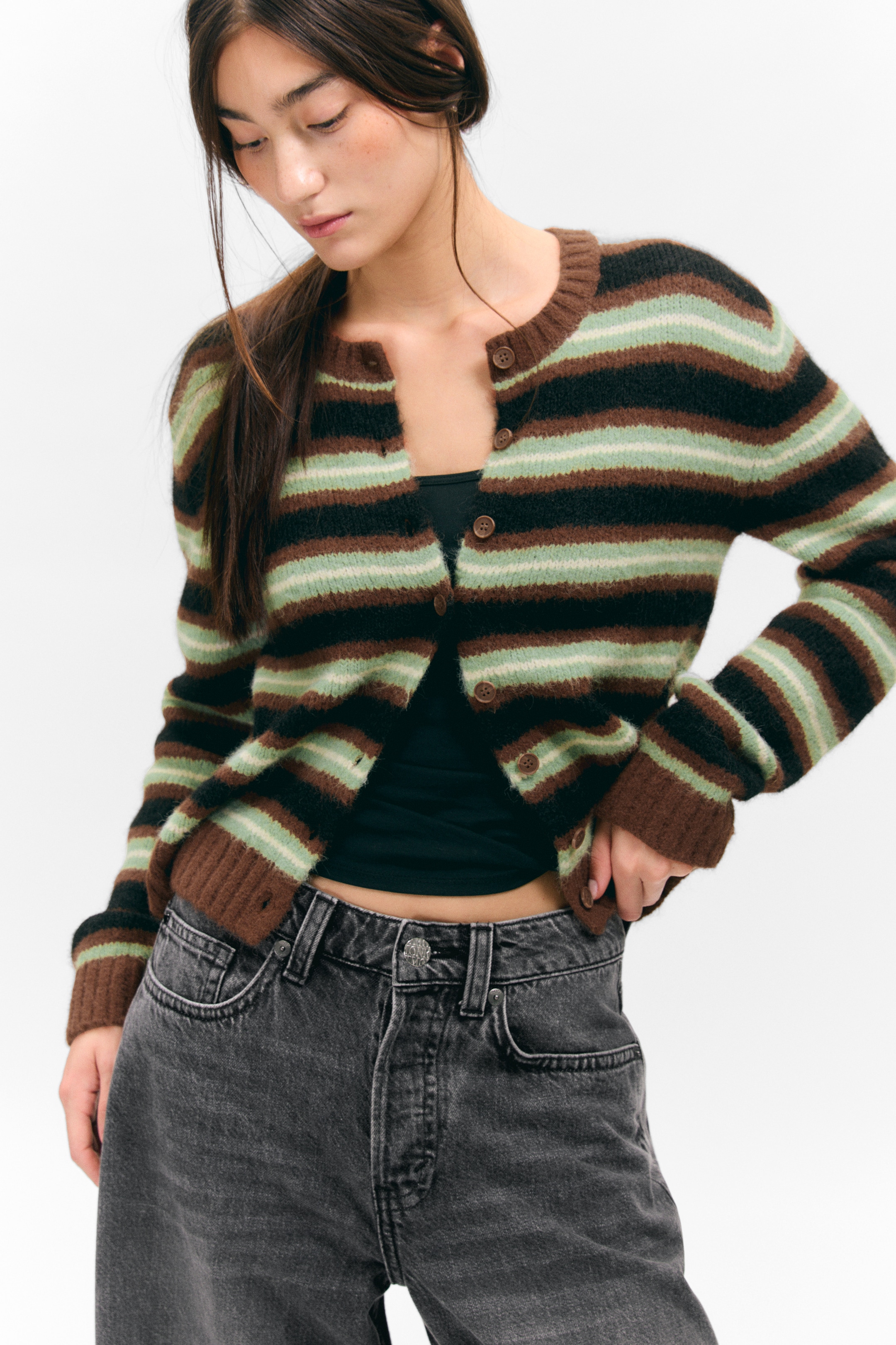 Ingrandisci l'immagine: Un cardigan a righe con bottoni sul davanti è indossato, leggermente inclinato a destra, sopra un top scuro. Presenta righe orizzontali in marrone, nero e verde chiaro, enfatizzate da righe bianche più sottili, e mostra uno scollo rotondo, maniche lunghe con polsini a coste e un orlo a coste, chiuso sul davanti con bottoni marroni.