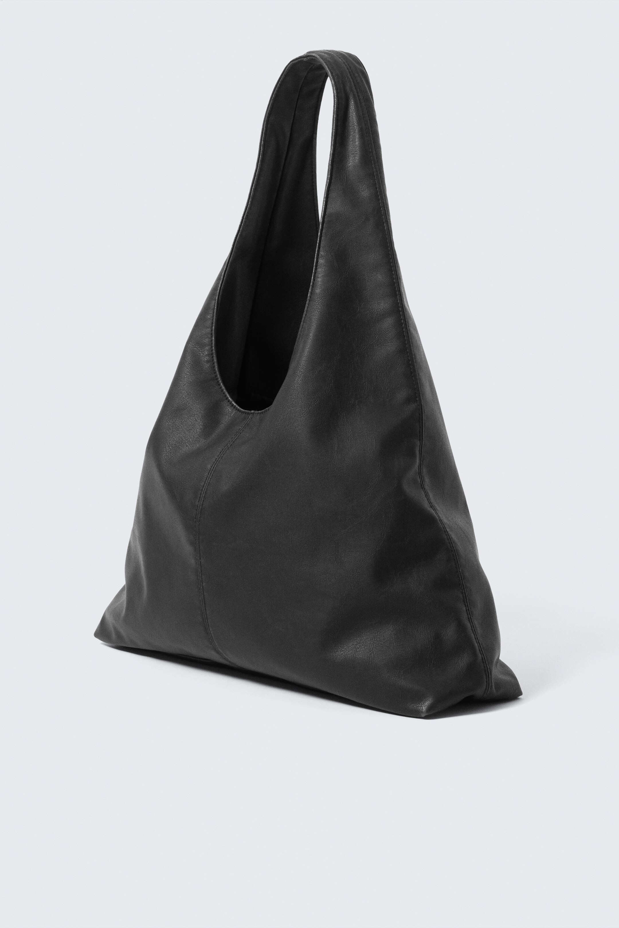Grösseres Bild ansehen: Eine schwarze Hobo-Tasche steht schräg, um ihre weiche, lässige Struktur zu zeigen. Sie präsentiert einen breiten, integrierten Riemen und eine subtile vertikale Naht, die an der Vorderseite verläuft.