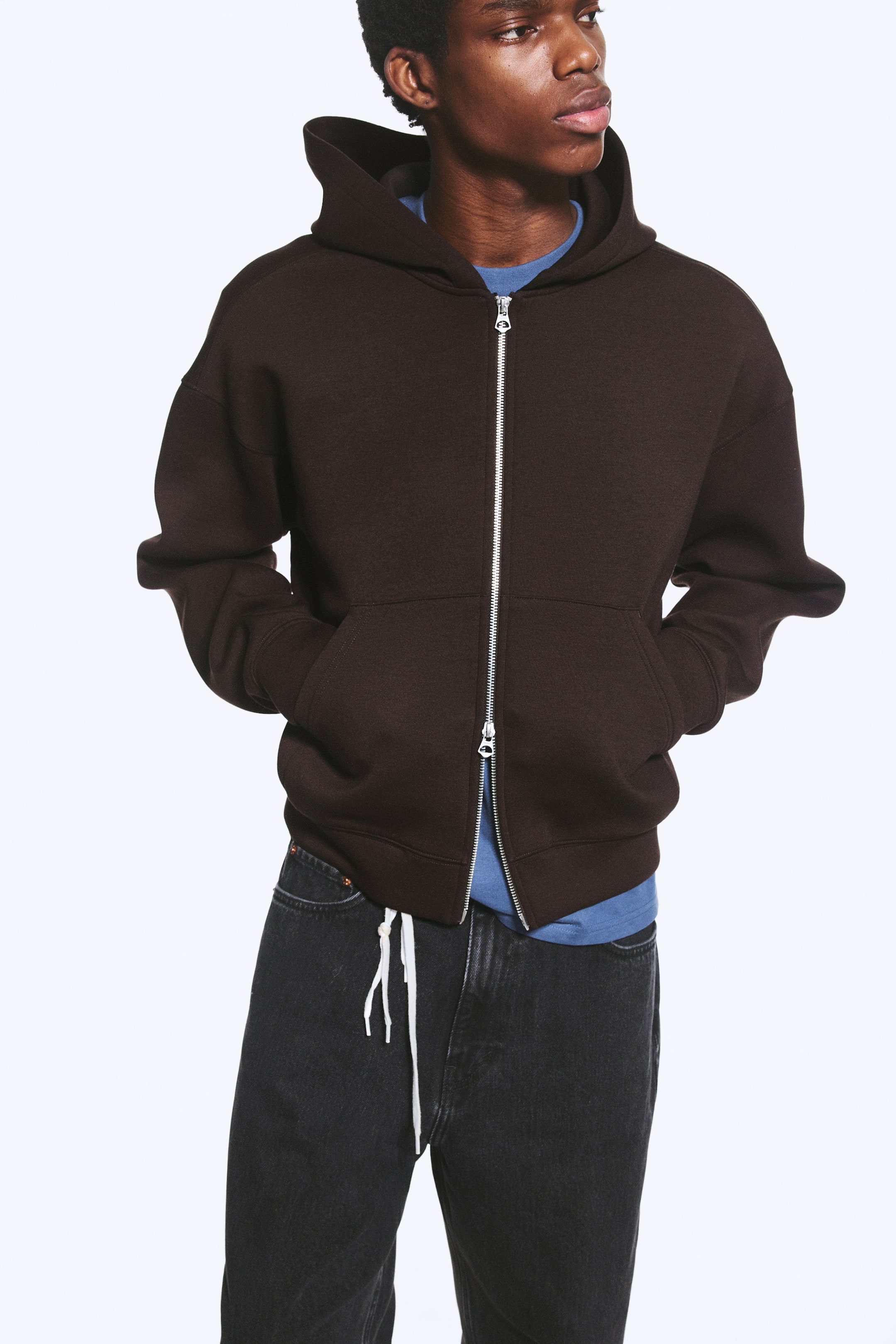 Ver imagen más grande: La modelo lleva puesta una sudadera con capucha de color marrón oscuro de corte regular, con hombros caídos, puños acanalados y un dobladillo acanalado. Su cremallera frontal plateada de doble sentido está parcialmente abierta desde abajo, y las manos de la modelo descansan en los bolsillos tipo canguro delanteros, dejando ver una camiseta azul debajo.