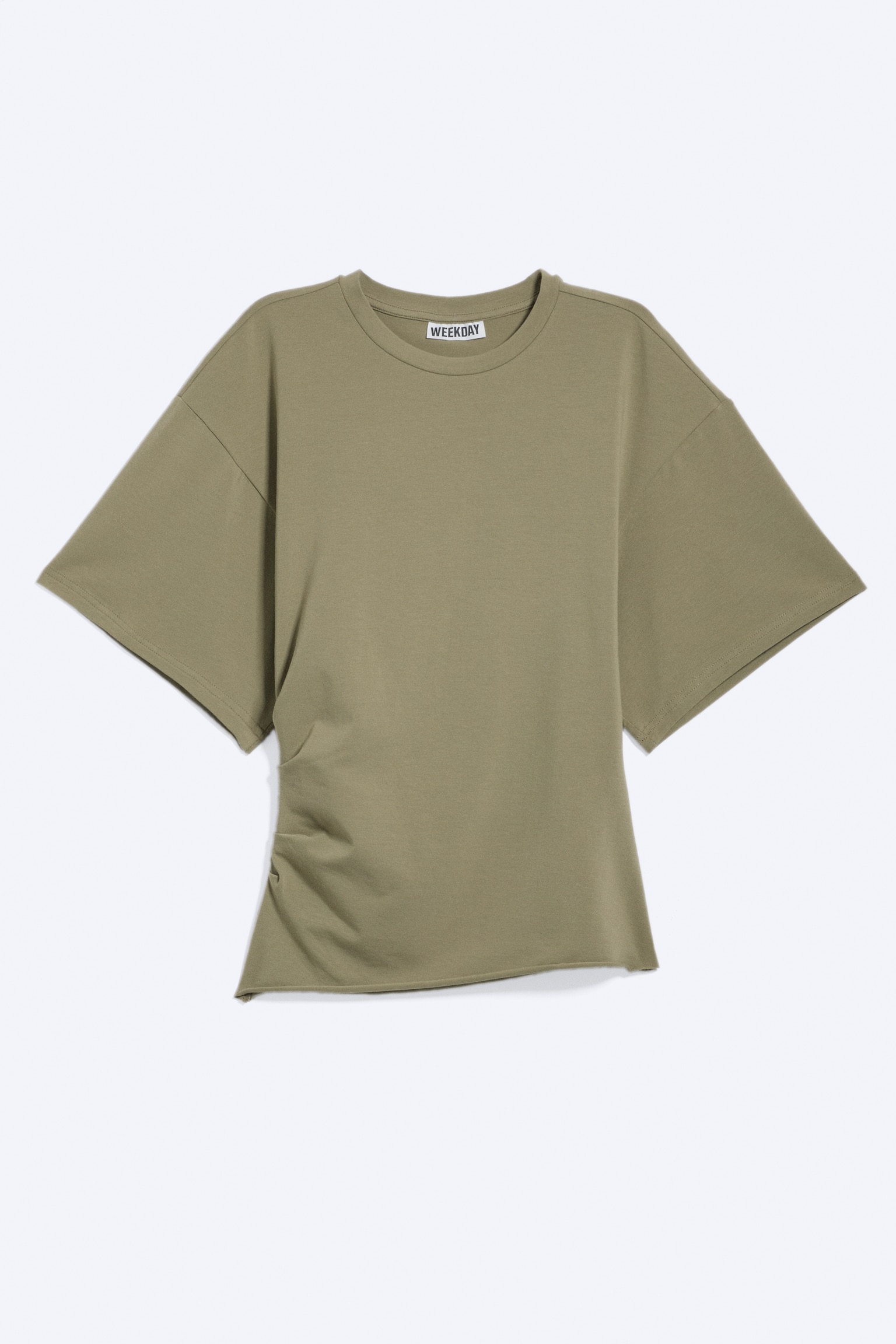 En T-shirt visar sin framsida, med en rund hals och boxiga korta ärmar. Den har placerade veck som skapar en draperad effekt på vänster sida och en rå kant längs nederkanten, med en 'WEEKDAY'-etikett synlig vid halslinningen.