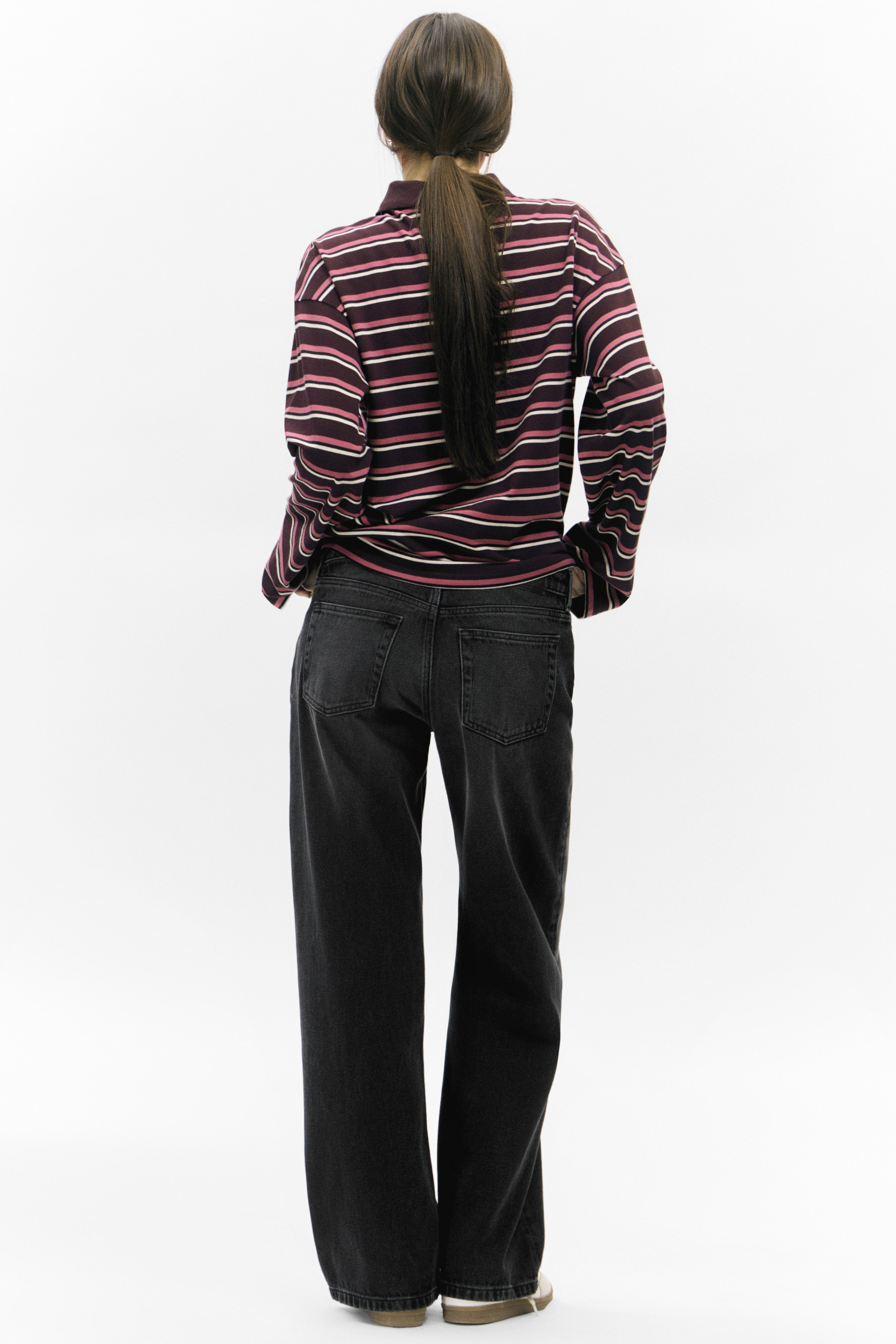 Visualizza immagine più grande: Un top polo a maniche lunghe a righe è indossato dalla modella, caratterizzato da un colletto polo a coste, spalle cadenti e una silhouette rilassata. L'orlo dritto del top appare leggermente tirato mentre la modella tiene le mani nelle tasche dei jeans scuri.
