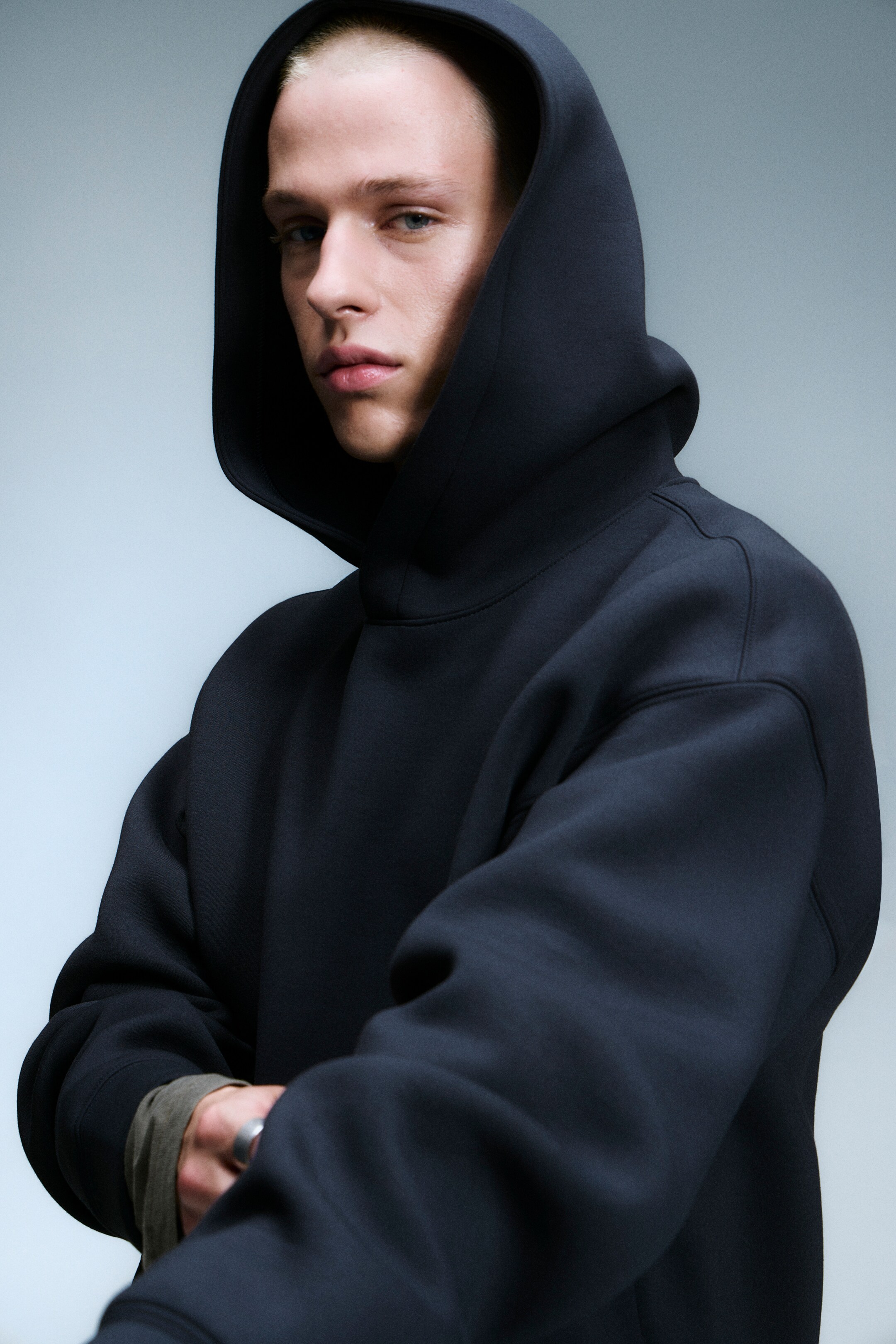 Grotere afbeelding bekijken: De zwarte pullover hoodie is voorzien van een grote, gestructureerde capuchon die het hoofd van het model bedekt. Het toont een gladde, stevige stof met verlaagde schouders en lange mouwen, en een gedempt groen lang mouwenvest is zichtbaar eronder en komt uit de manchetten.