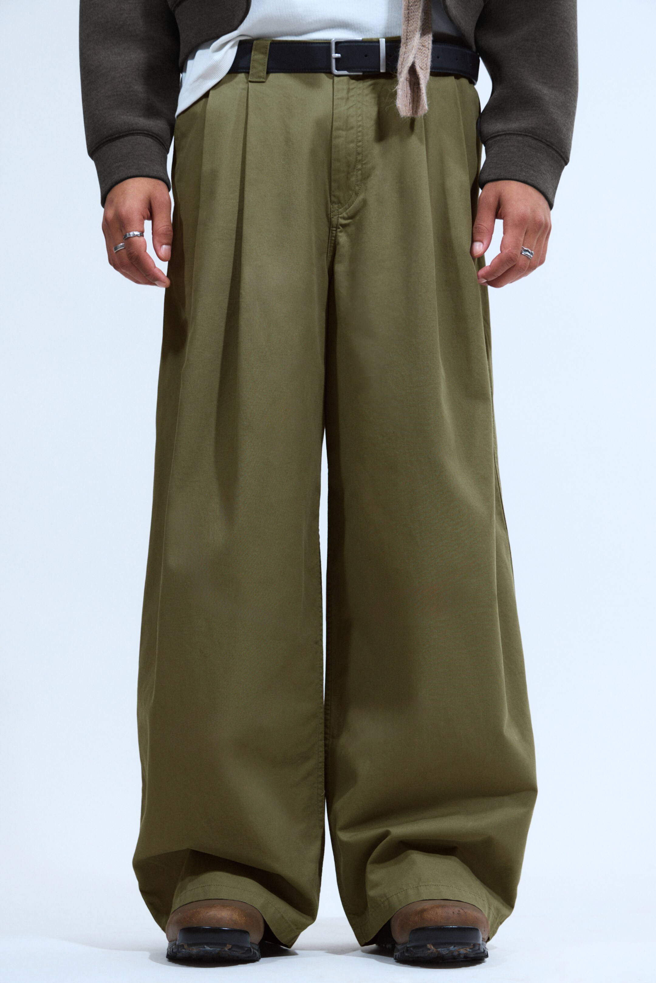 Agrandir l'image: Les chinos olive verts surdimensionnés orientés vers l'avant présentent une silhouette ample et une jambe large, avec des plis avant proéminents, des poches latérales en biais et des passants de ceinture tenant une ceinture noire. Une fermeture à bouton supérieur est visible, et le tissu apparaît comme un sergé de coton.
