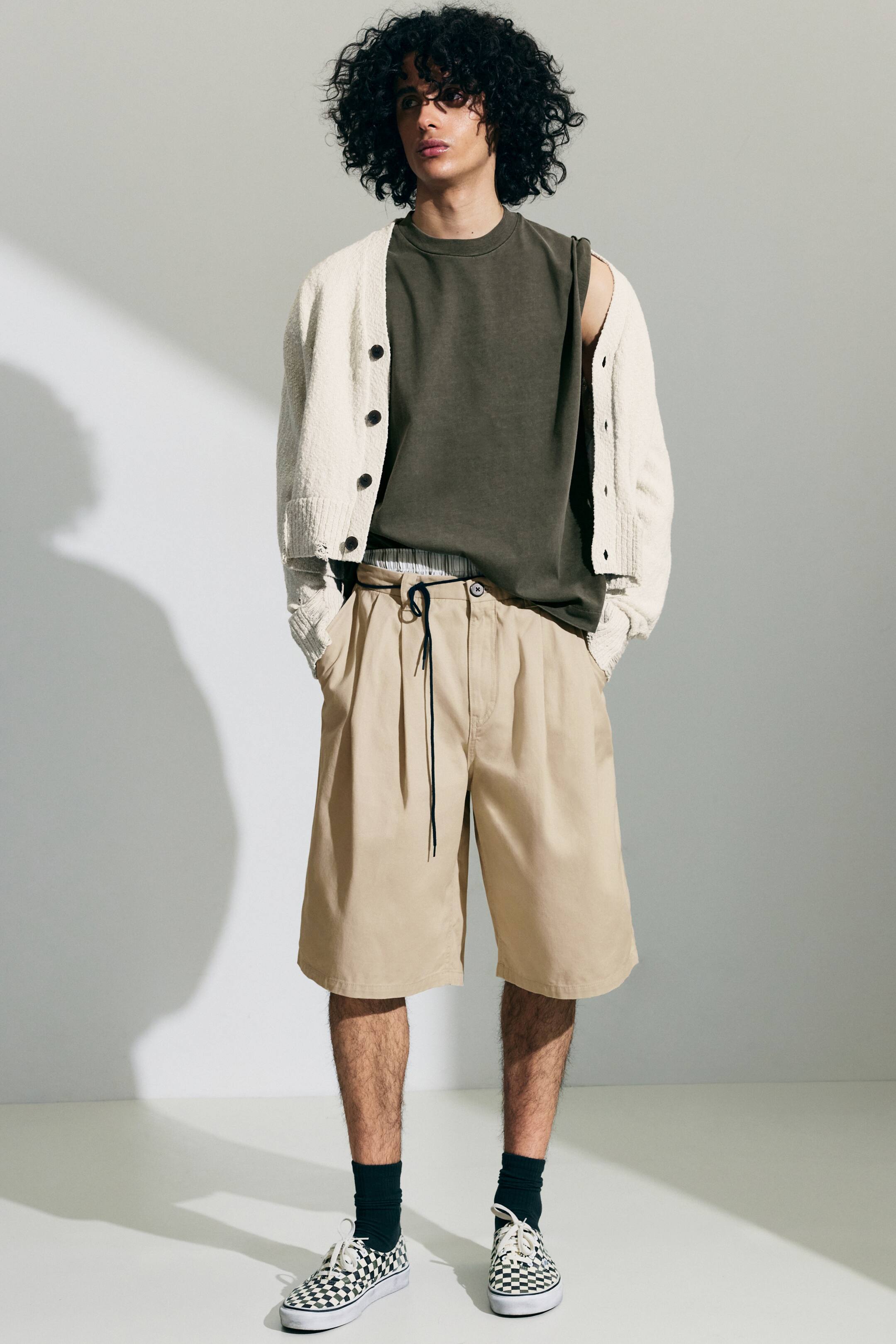 Visualizza immagine più grande: Un outfit presenta un cardigan in maglia testurizzata color crema, indossato aperto su un top verde oliva senza maniche con scollo a giro e apertura delle maniche effetto usurato, abbinato a shorts khaki a gamba larga con pieghe e vita con coulisse nera. L'ensemble è completato da calzini scuri e sneakers slip-on a scacchi bianche e nere.
