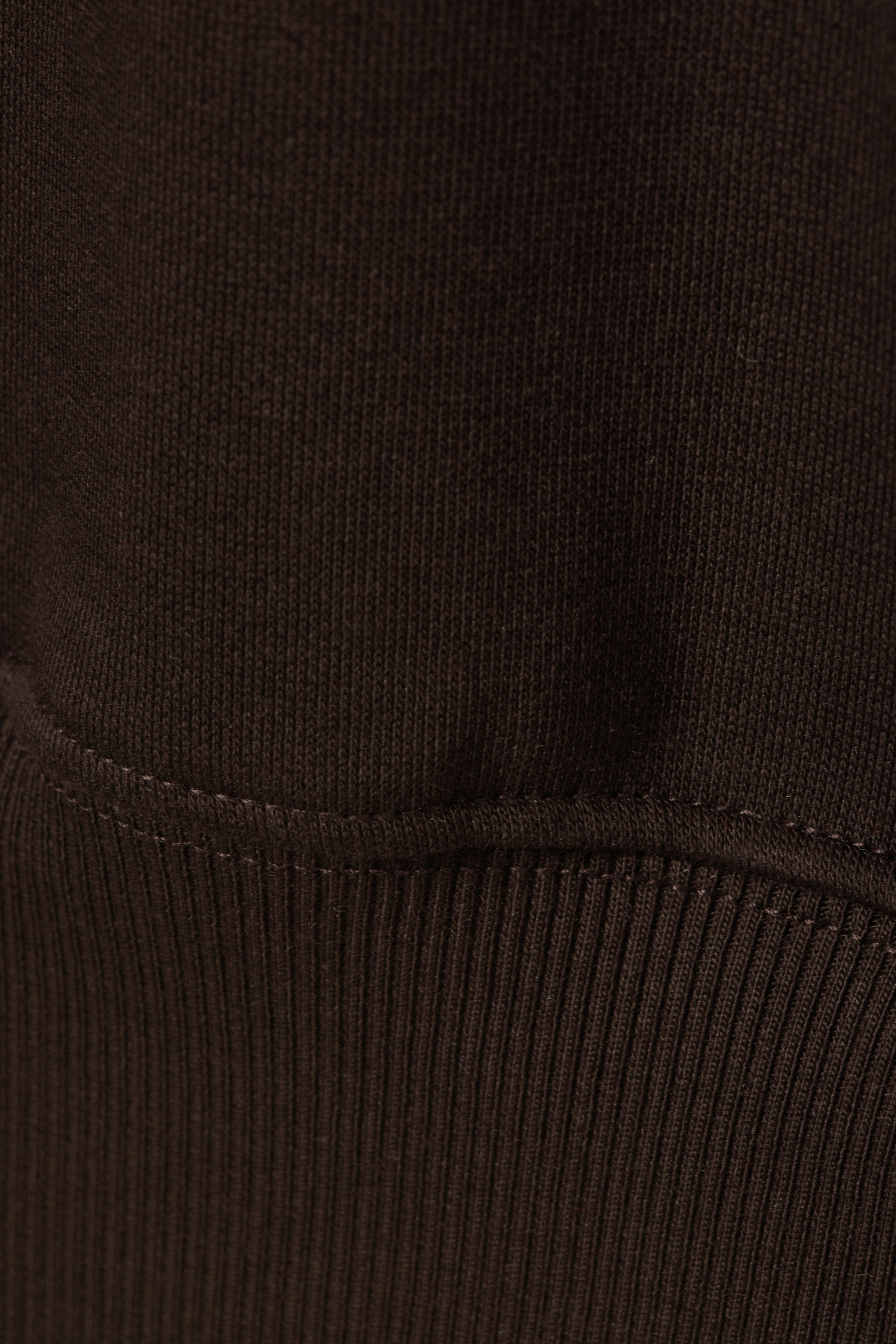 Agrandir l'image: Un tissu brun foncé présente une texture de tricot lisse dans la partie supérieure. Une ligne de couture avec des points visibles sépare cela d'une section en tricot côtelé en dessous, suggérant un poignet ou un ourlet.