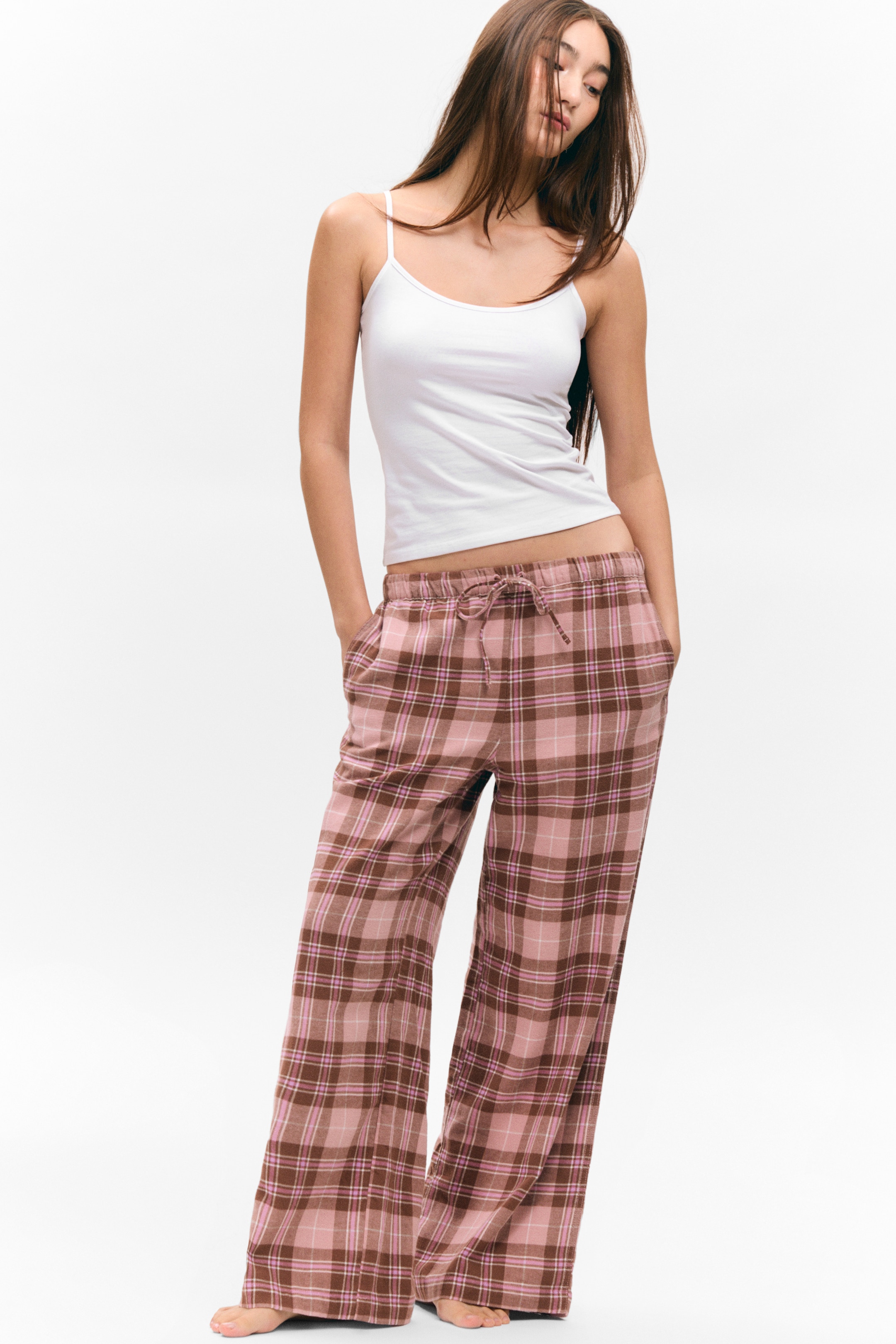 View larger image: Wide-leg plaid pants are being presented from a front-facing angle, featuring a pink and brown plaid pattern, a drawstring waist, and side pockets. They are being styled with a white camisole, which is featuring thin spaghetti straps.
