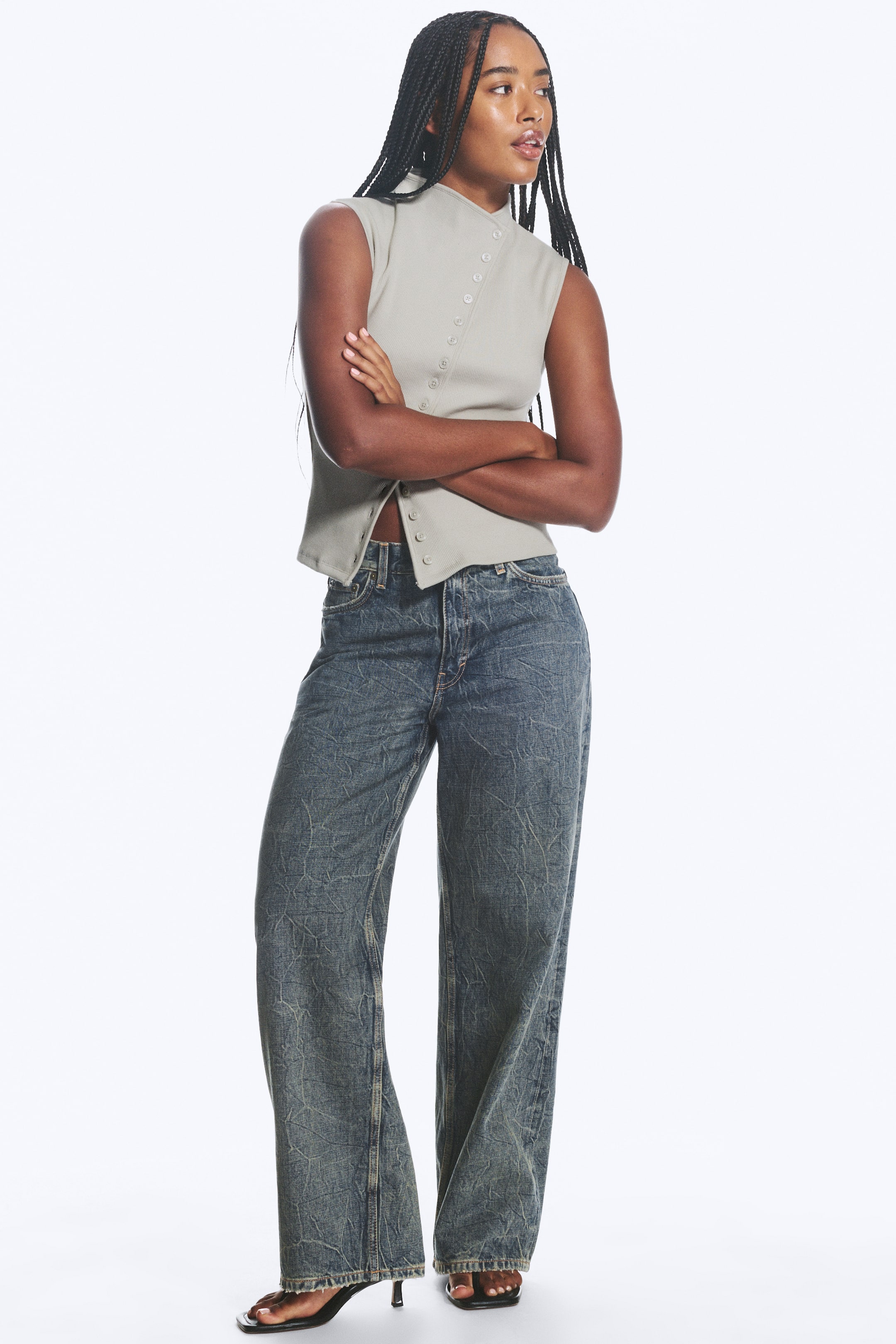 View larger image: The pale green sleeveless top features a high stand collar and an asymmetrical button placket with visible buttons running diagonally across the front, with some buttons undone at the hem. The top and the dark wash blue jeans, which showcase a crinkled texture and a wide-leg design, are both presented facing slightly to the right.