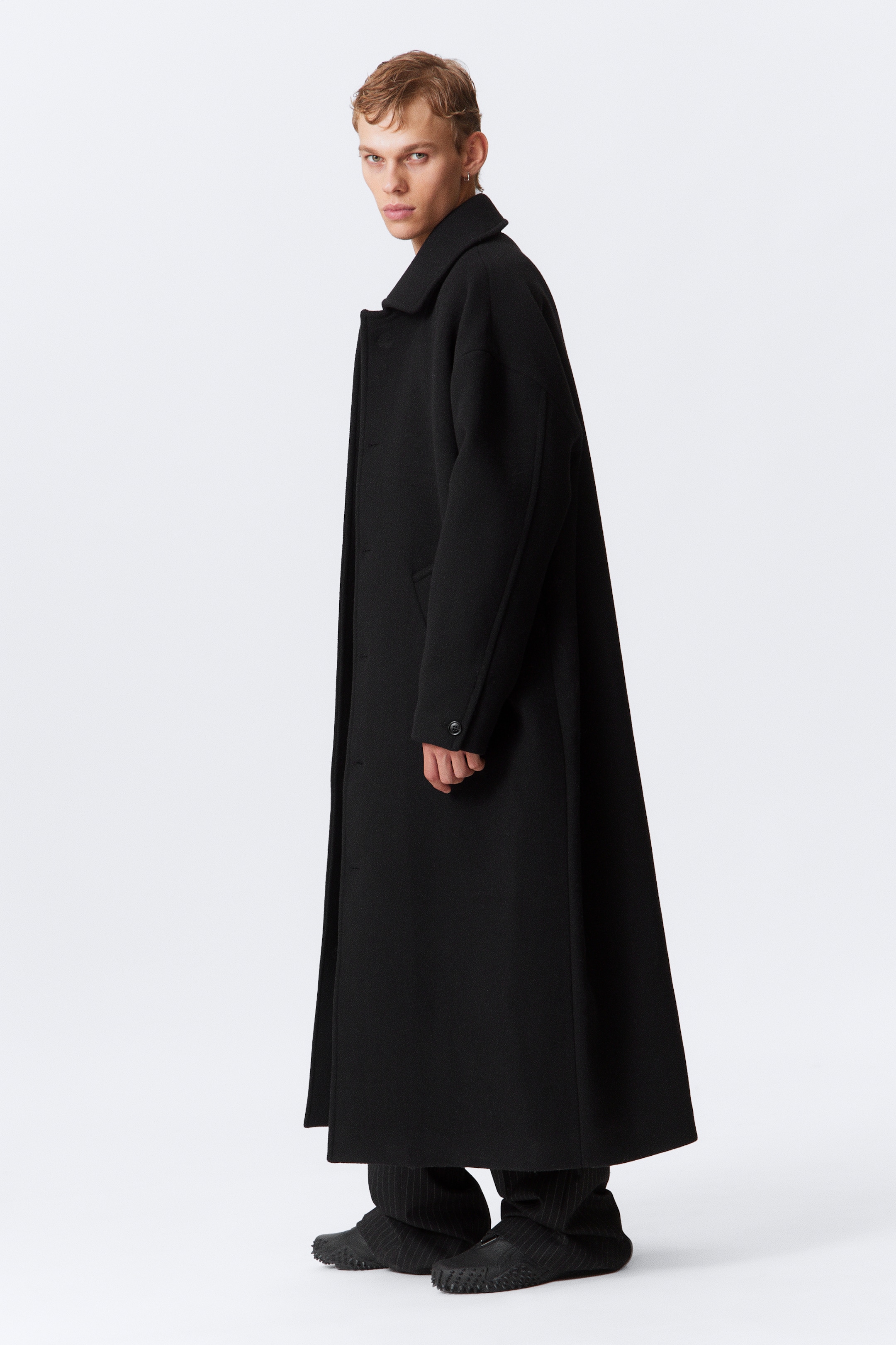 Größeres Bild anzeigen: Ein langer, schwarzer Mantel mit übergroßer Silhouette verfügt über einen Umlegekragen, einen Knopfverschluss vorne, schräge Leistentaschen und zweiteilige Ärmel. Das Model trägt den Mantel, der über gestreiften Hosen geschichtet ist.