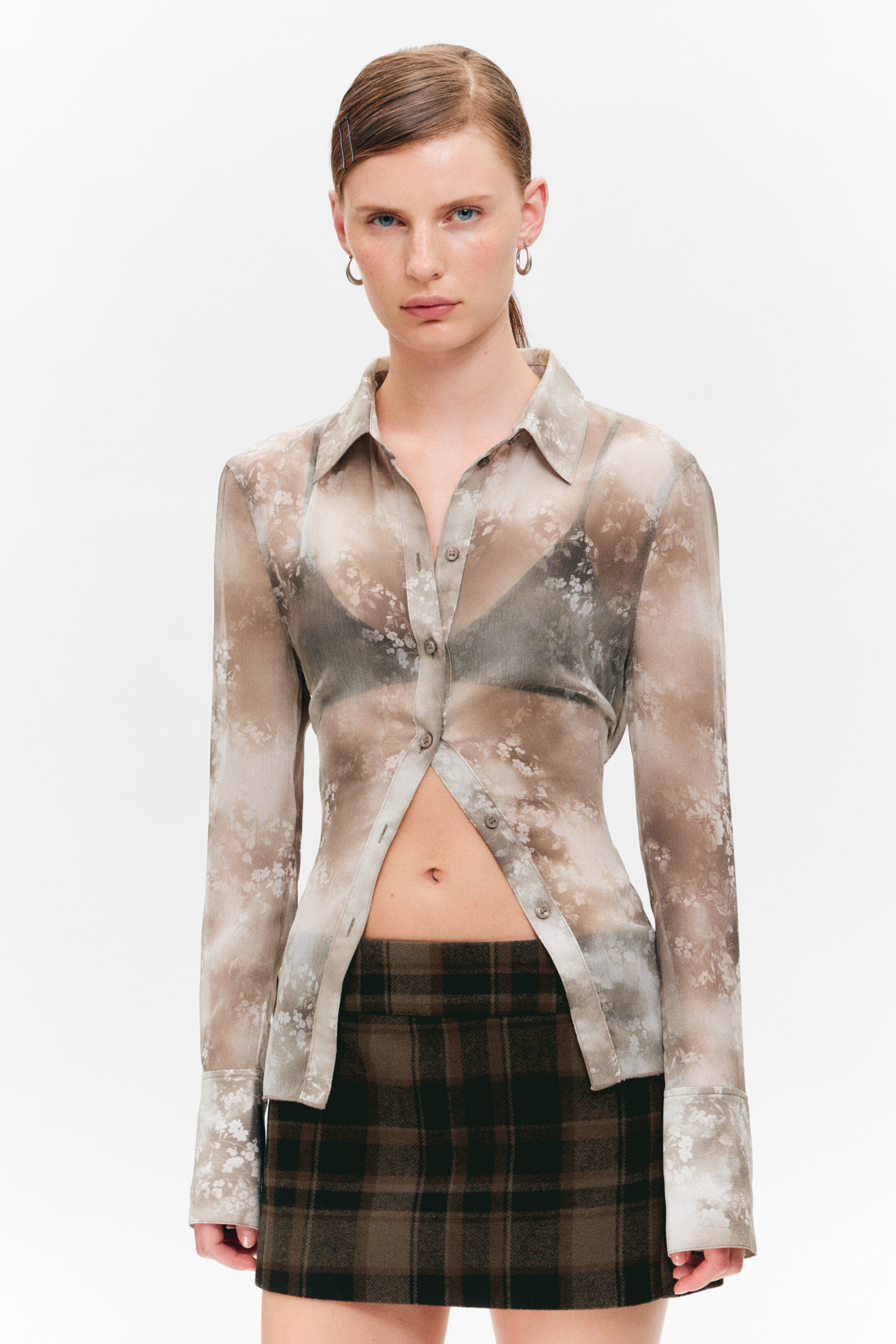 View larger image: A sheer, collared long-sleeve shirt with buttoned cuffs and a light brown floral pattern is being worn, partially unbuttoned to reveal a dark bra underneath and the midriff. It is layered over a brown and dark brown plaid mini skirt.