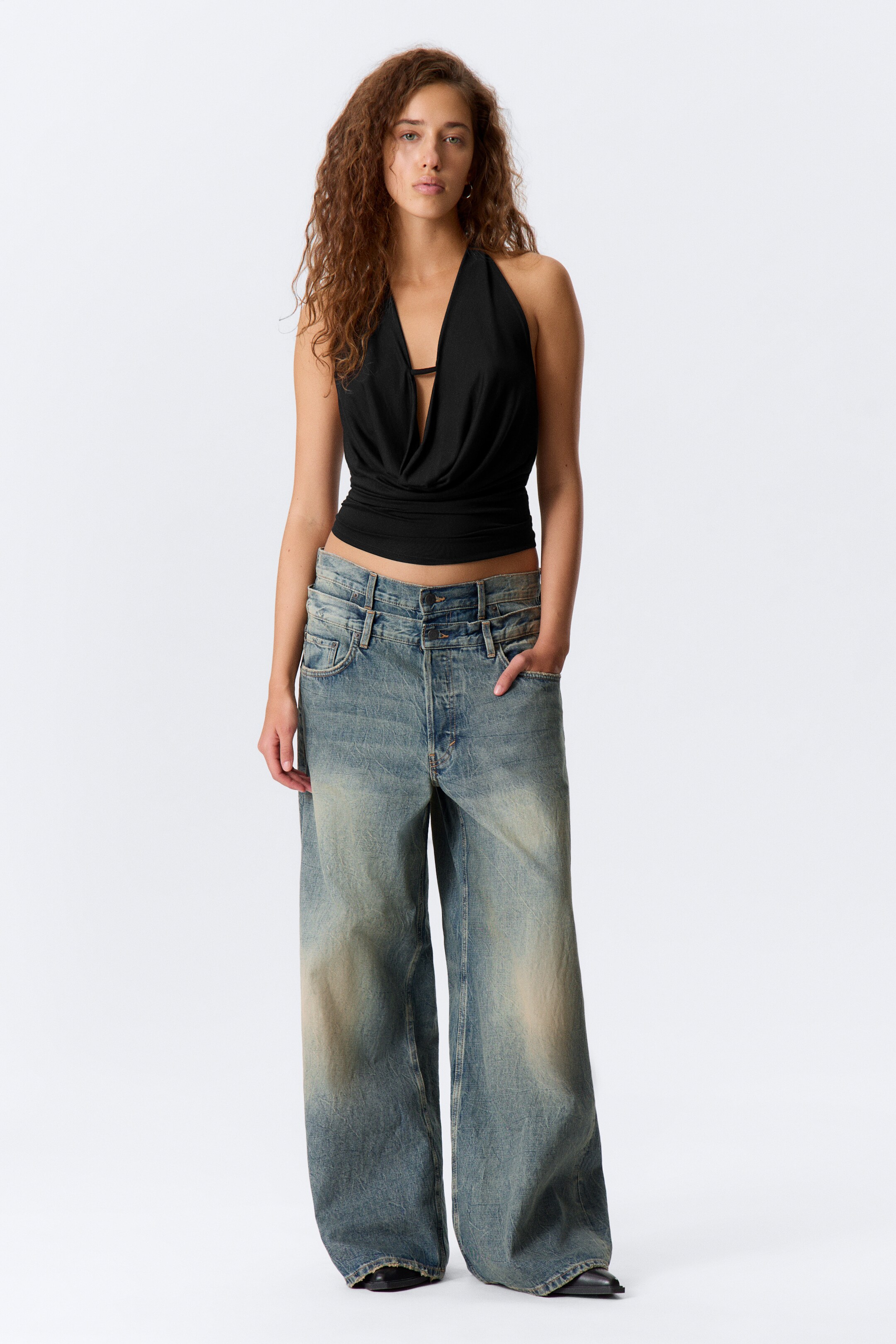 Grotere afbeelding bekijken: Een zwarte haltertop wordt gekenmerkt door een draperende cowl halslijn en een cropped zoom, die een kleine band eronder onthult. Het wordt frontaal gepresenteerd, gecombineerd met lichtblauwe denim jeans met wijde pijpen die een dubbele tailleband met meerdere knopen en een vervaagde, versleten afwerking tonen.
