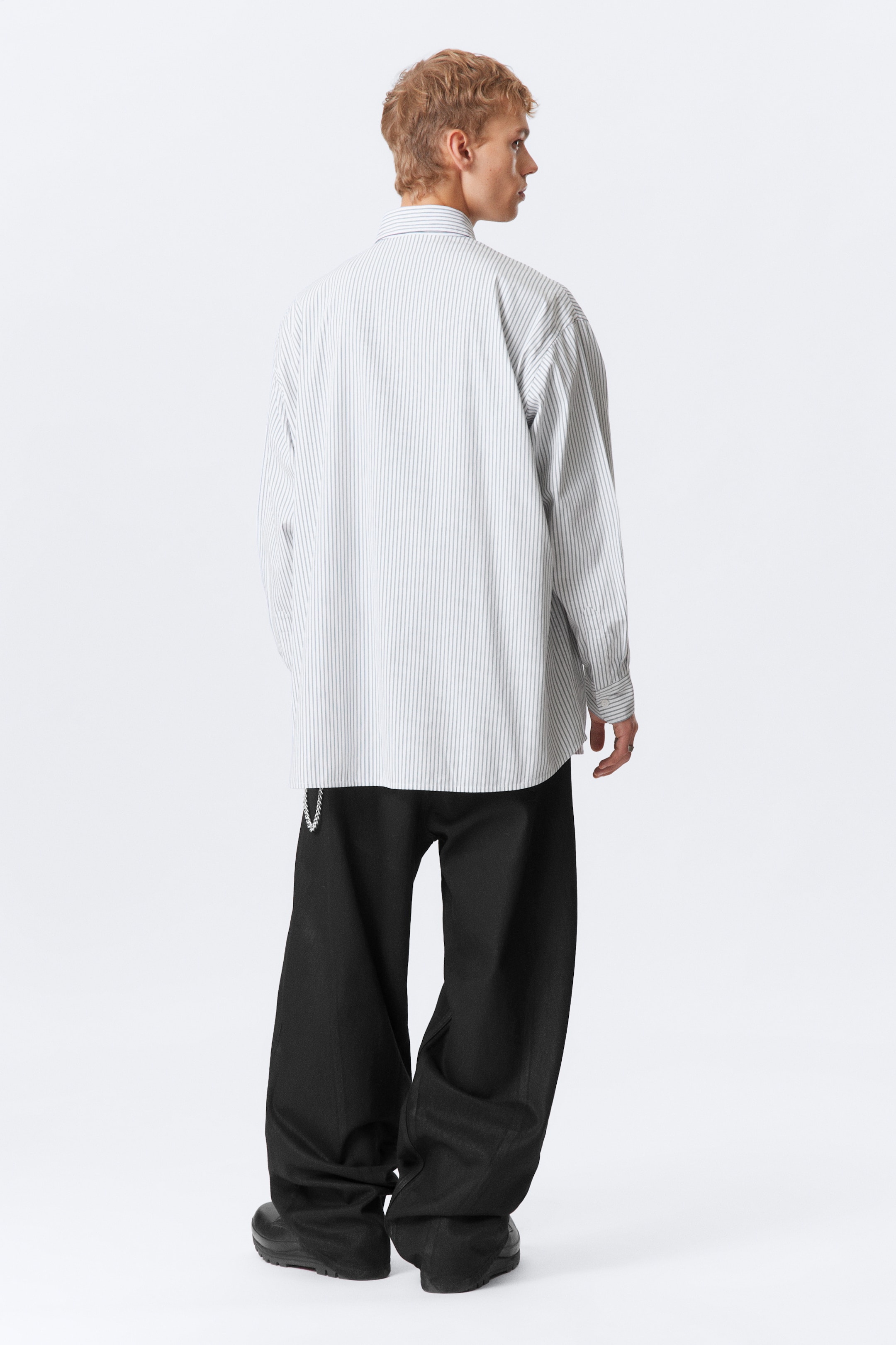 View larger image: An oversized white shirt with thin vertical grey stripes is being worn, seen from the back, featuring long sleeves and a visible collar. Black wide-leg trousers are being paired with the shirt, fitting loosely and pooling over black chunky shoes, with a silver chain hanging from the waistband.