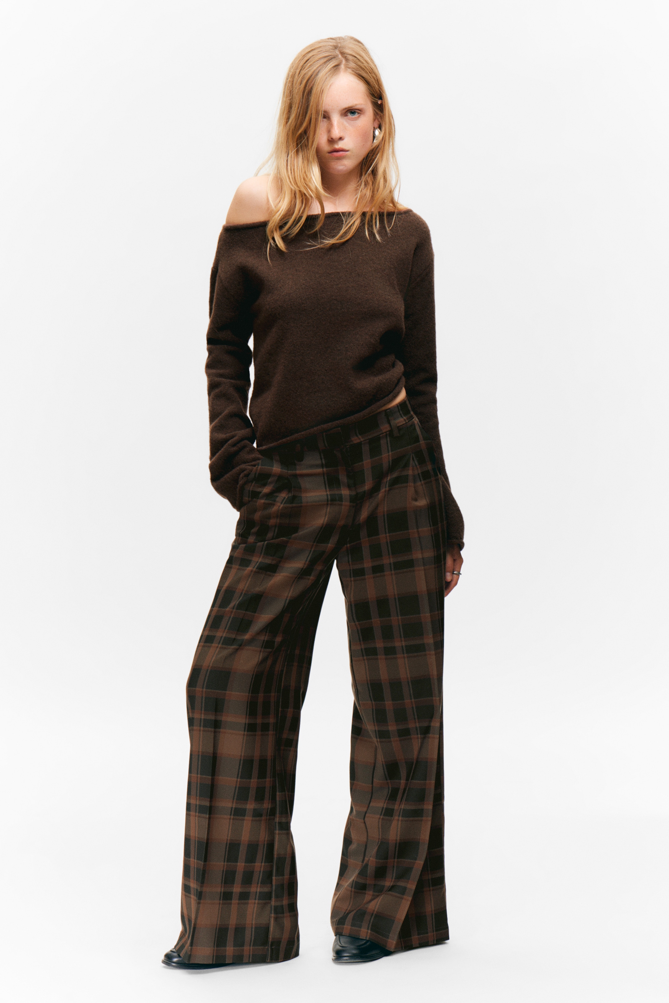 Grösseres Bild ansehen: Ein dunkelbrauner Off-Shoulder-Pullover wird präsentiert, der einen breiten Ausschnitt aufweist, der eine Schulter freilegt, und lange Ärmel hat. Er wird mit weit geschnittenen Hosen kombiniert, die ein braun-schwarzes Karomuster mit rötlich-braunen Akzenten zeigen und vorne Falten aufweisen.