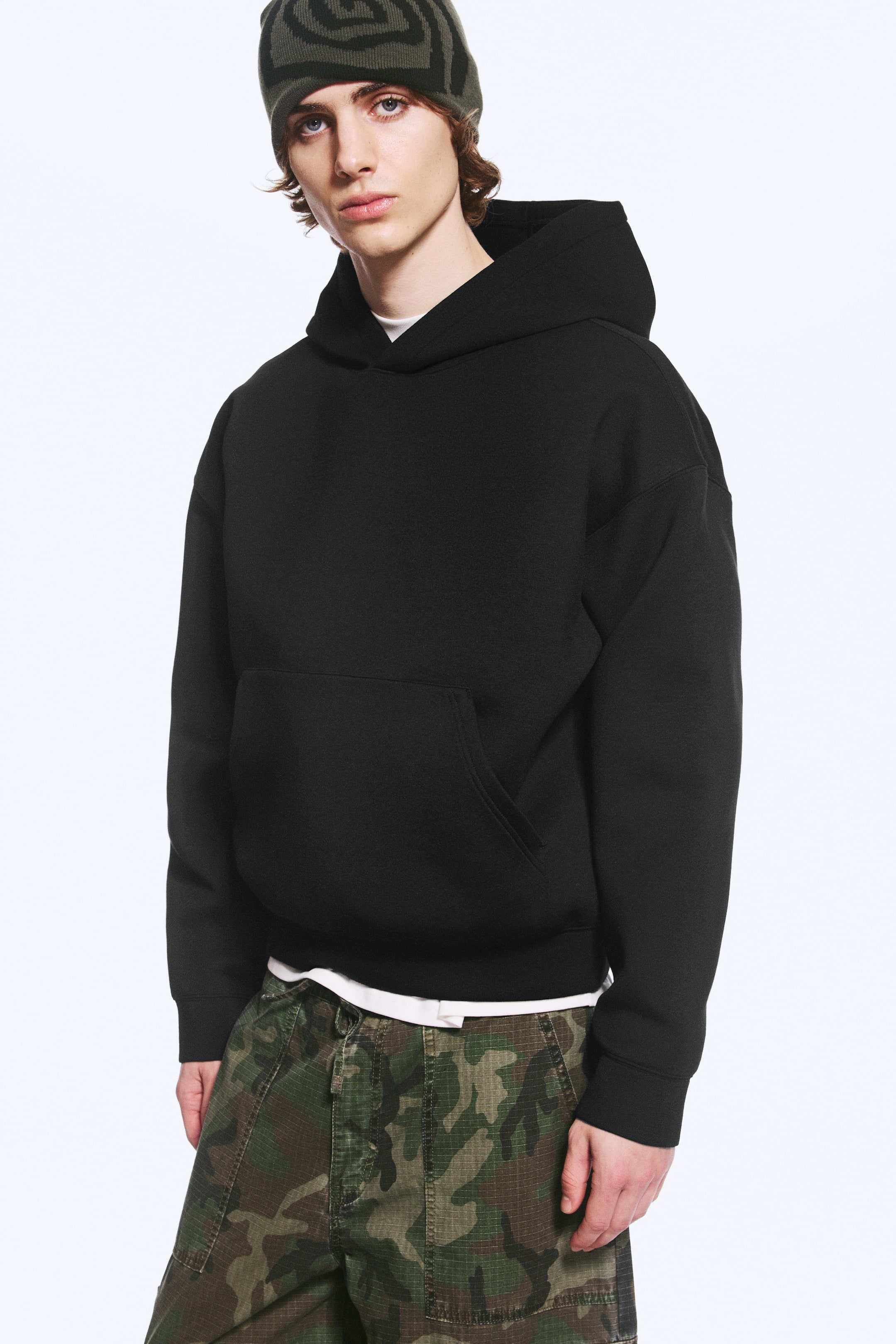 Visa större bild: En svart hoodie i lös passform i ett strukturerat scuba-tyg bärs av modellen. Hoodien har nedhasade axlar, en känguruficka framtill och släta muddar, och är lagerlagd över en vit T-shirt och matchas med camouflagebyxor.