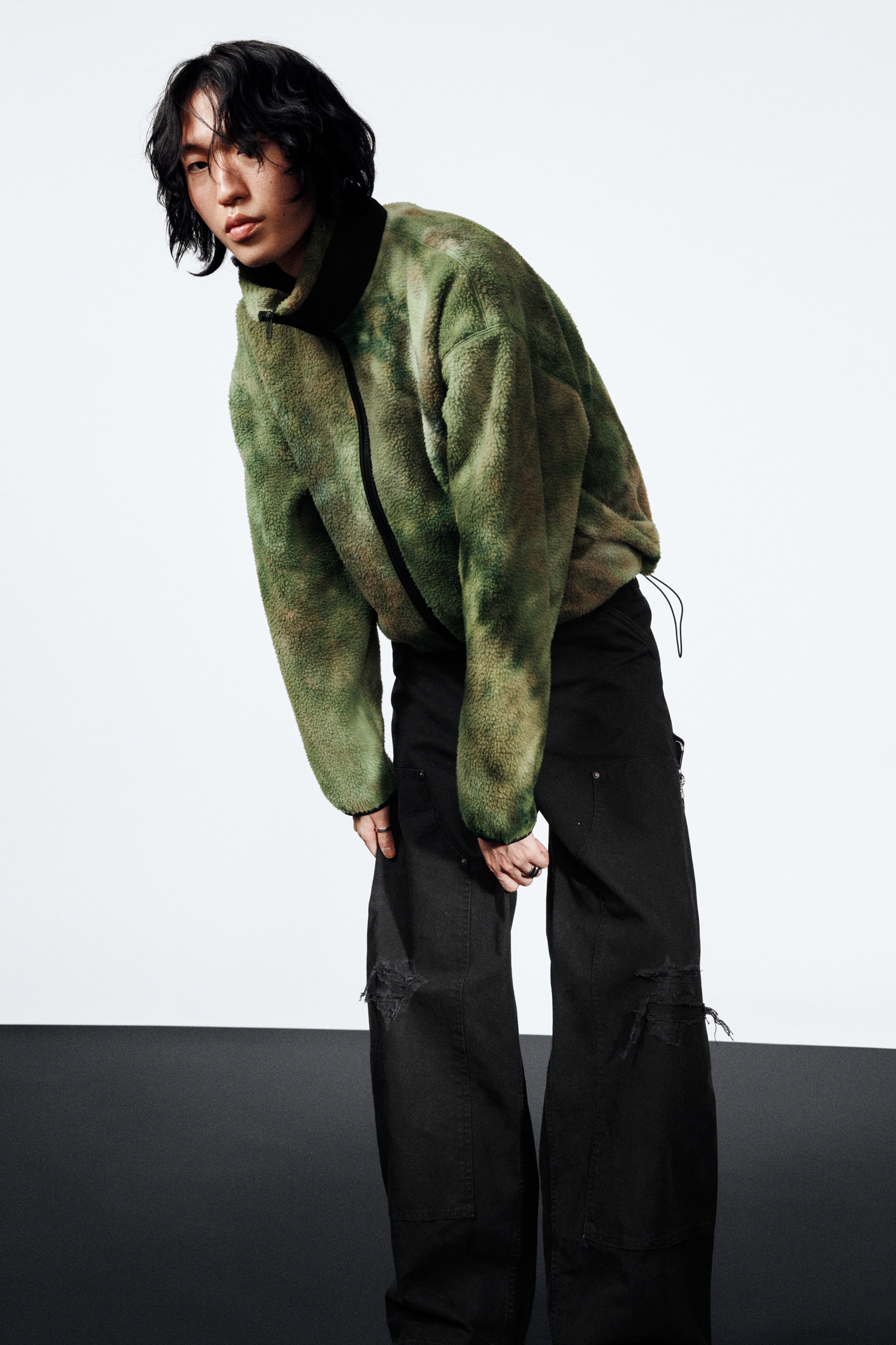 Grösseres Bild ansehen: Eine grün-braun batikgemusterte Fleecejacke wird getragen, die über einen schwarzen hohen Kragen, einen zentralen schwarzen Reißverschluss, einen Kordelzug am Saum und elastische Bündchen verfügt. Sie wird mit schwarzen, weit geschnittenen Hosen kombiniert, die an den Knien abgenutzt sind.
