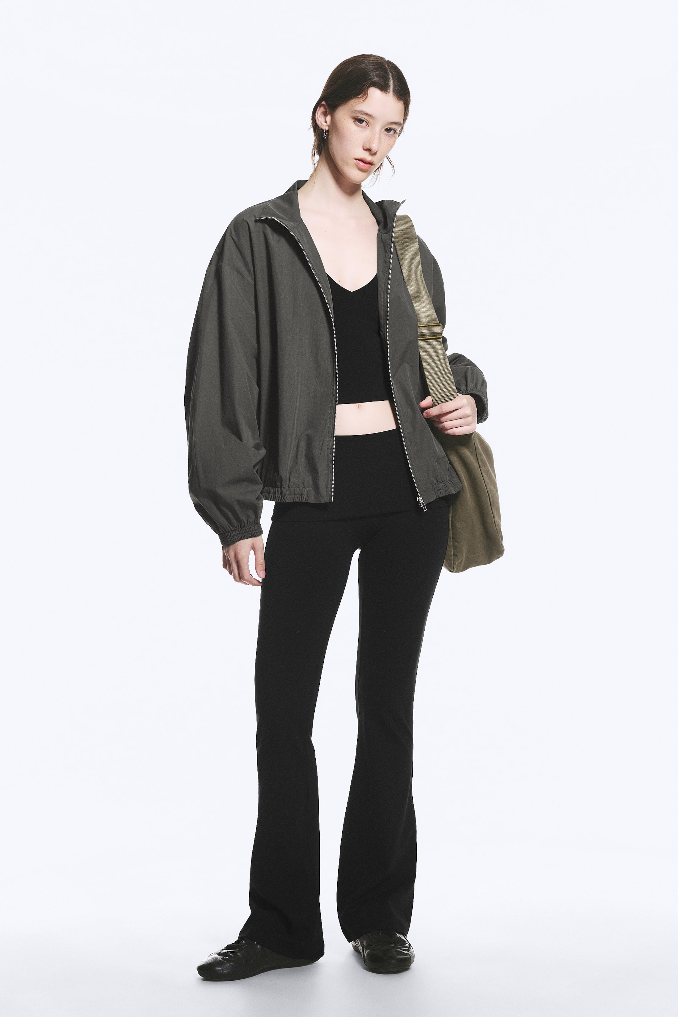 Se større billede: En mørkegrå bomberjakke med stand-up krave, en fuld frontlynlås, voluminøse lange ærmer samlet ved manchetterne og en elastisk kant. Den har en afslappet pasform og bæres åbent over en sort crop top.