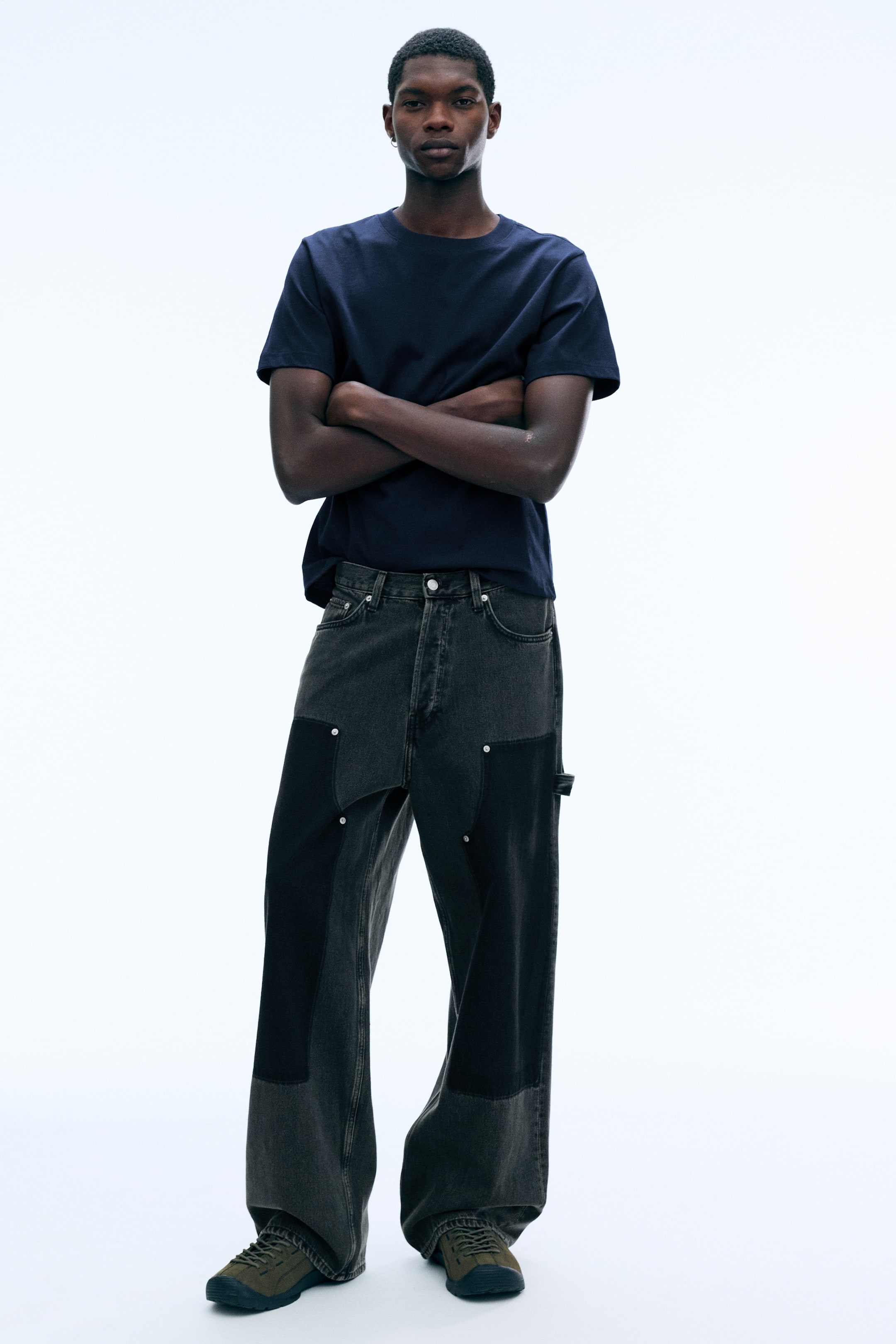 Agrandir l'image: Une paire de jeans baggy à coupe ample en denim gris foncé, avec des empiècements noirs contrastants au niveau des cuisses et une boucle porte-marteau, est portée par le mannequin. Les jeans sont associés à un t-shirt bleu marine rentré à l’intérieur et à des sneakers vert foncé.