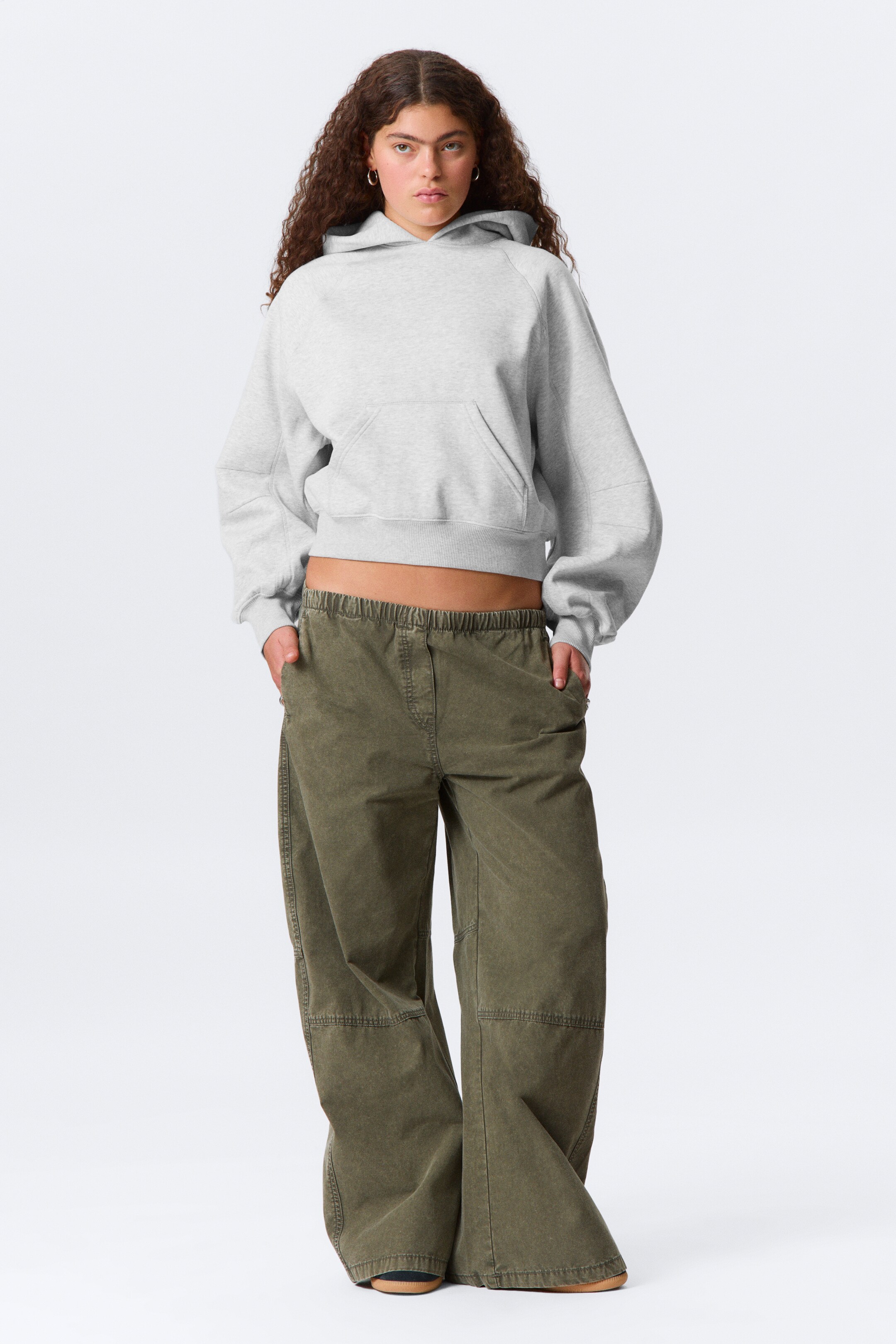 Visualizza immagine più grande: Una felpa corta grigia chiara, caratterizzata da un cappuccio, maniche raglan, polsini e orlo a coste, e una tasca a marsupio frontale, viene presentata con pantaloni ampi a gamba larga verde oliva. I pantaloni mostrano una cintura elasticizzata e cuciture a pannello sulle ginocchia, con entrambi i capi rivolti in avanti.