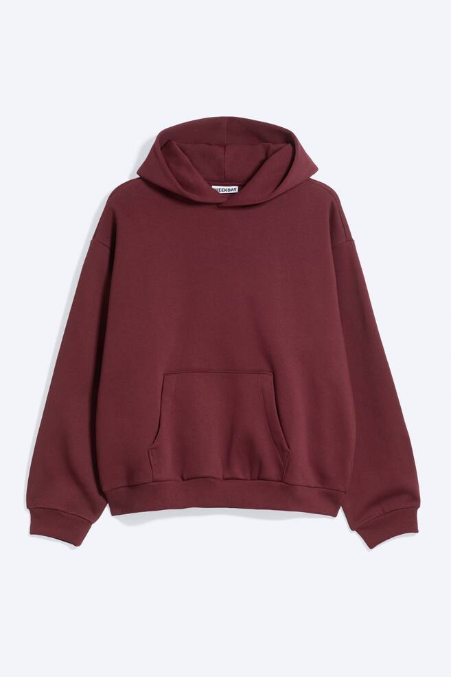 En burgundyfÃ¤rgad fleecehoodie Ã¤r vÃ¤nd framÃ¥t och visar en lÃ¶s, mjukt strukturerad passform med nedhasade axlar, en vikt huva och en rymlig kÃ¤nguruficka. Den har ribbade muddar och en ribbad fÃ¥ll med subtil rynkning som skapar en avslappnad, rundad siluett. - 1