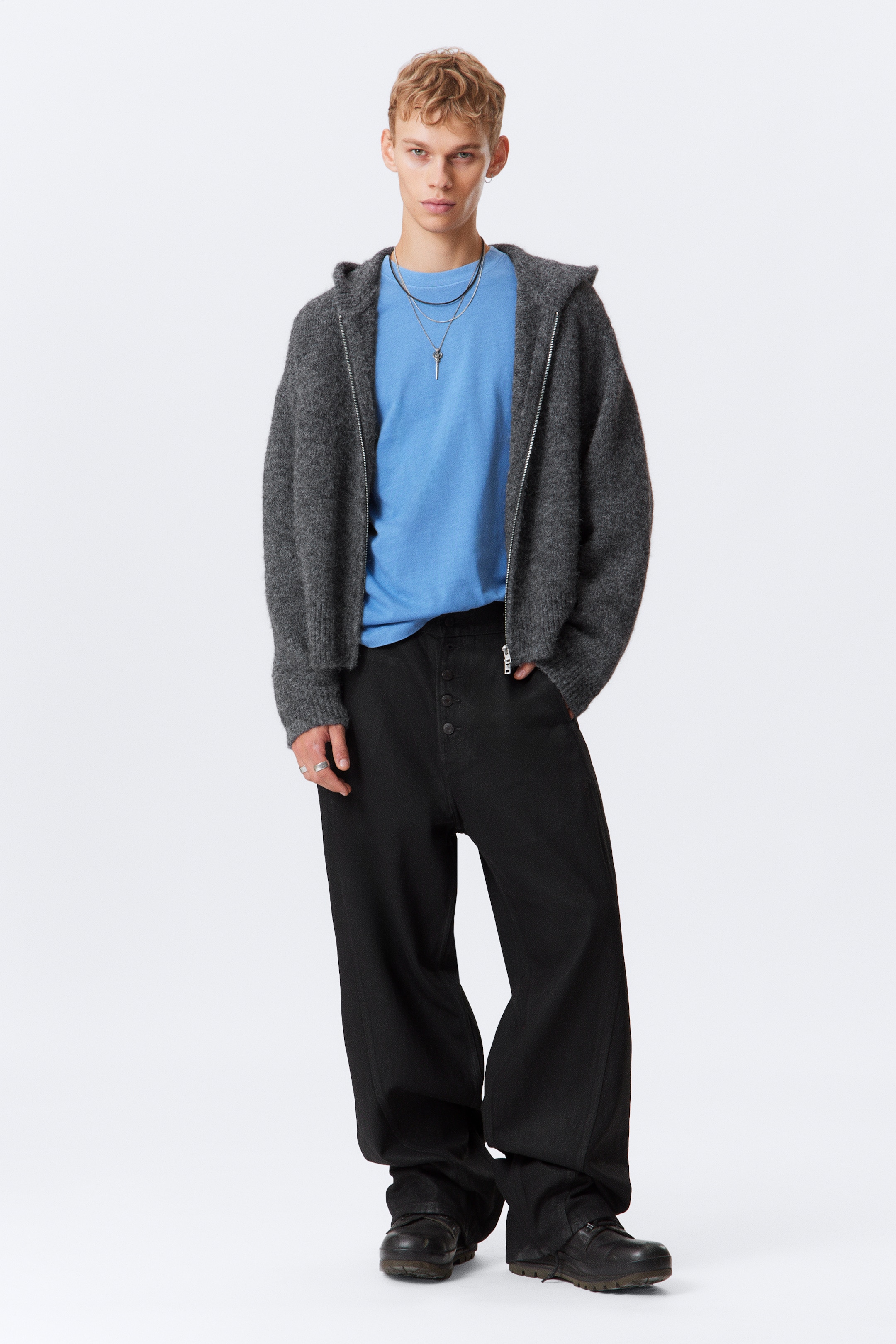 View larger image: A dark gray wool-blend zip-up hoodie with a loose, boxy fit is being worn by the model, featuring a visible two-way zip, ribbed cuffs, and a thick hood. The model is wearing the hoodie unzipped over a light blue t-shirt, revealing two necklaces, with their right hand resting in a front pocket.