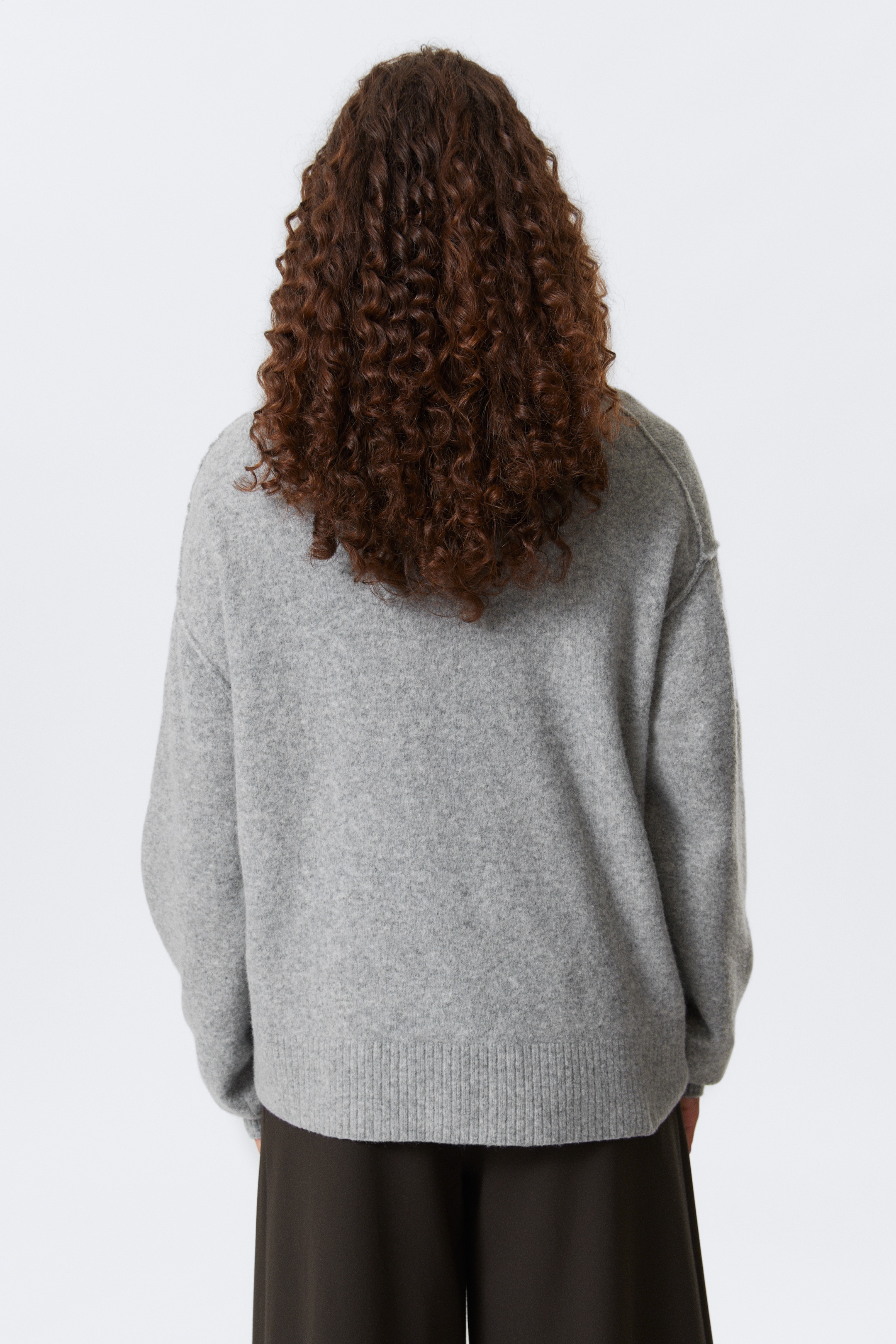 Größeres Bild ansehen: Ein hellgrauer Pullover wird von hinten betrachtet, mit einer entspannten Passform und abgesenkten Schulternähten. Er präsentiert einen breiten, gerippten Saum.