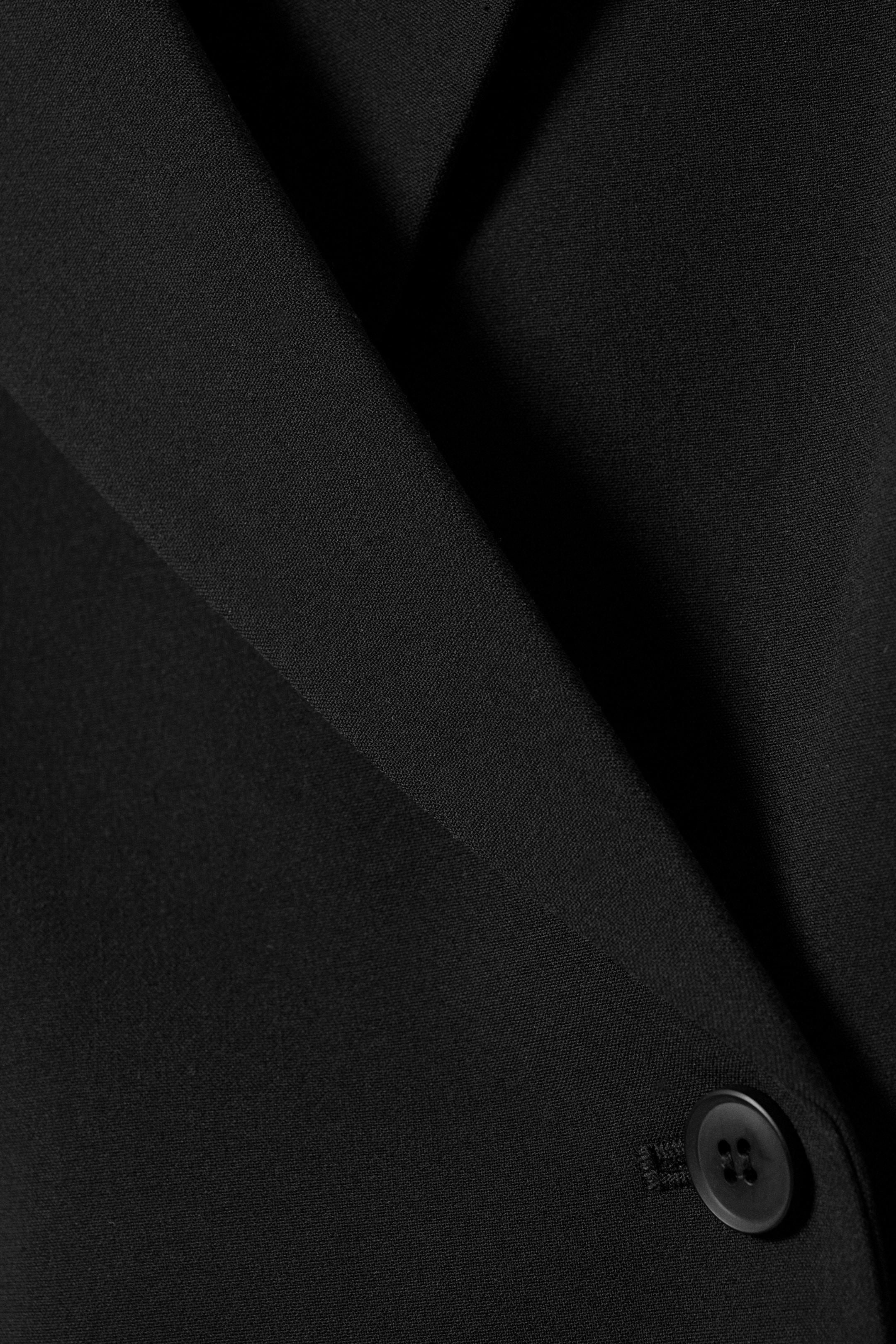 Visa större bild: En svart blazer visas i närbild, med en slät tygstruktur. En enda svart knapp med fyra hål är delvis synlig till höger, säkrad av en knapphålsögla, och en bred krage viks diagonalt över plagget.
