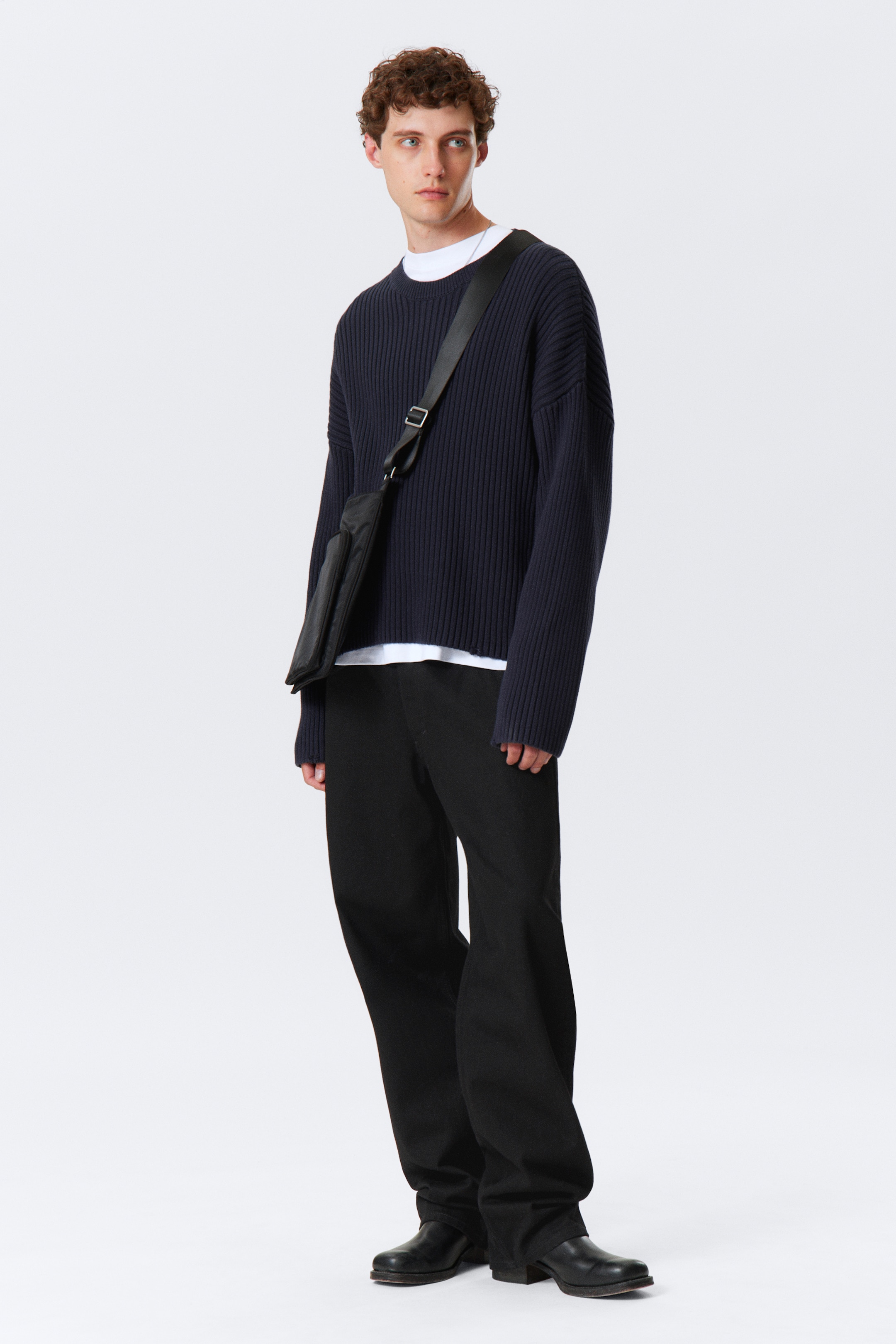 Visualizza immagine più grande: Un maglione a coste blu navy viene abbinato a pantaloni neri a gamba larga e una borsa a tracolla nera. Il maglione oversize presenta spalle scese, un girocollo, polsini a coste e un orlo dritto, con una t-shirt bianca stratificata sotto. I pantaloni a vestibilità rilassata si appoggiano su scarpe in pelle nera.
