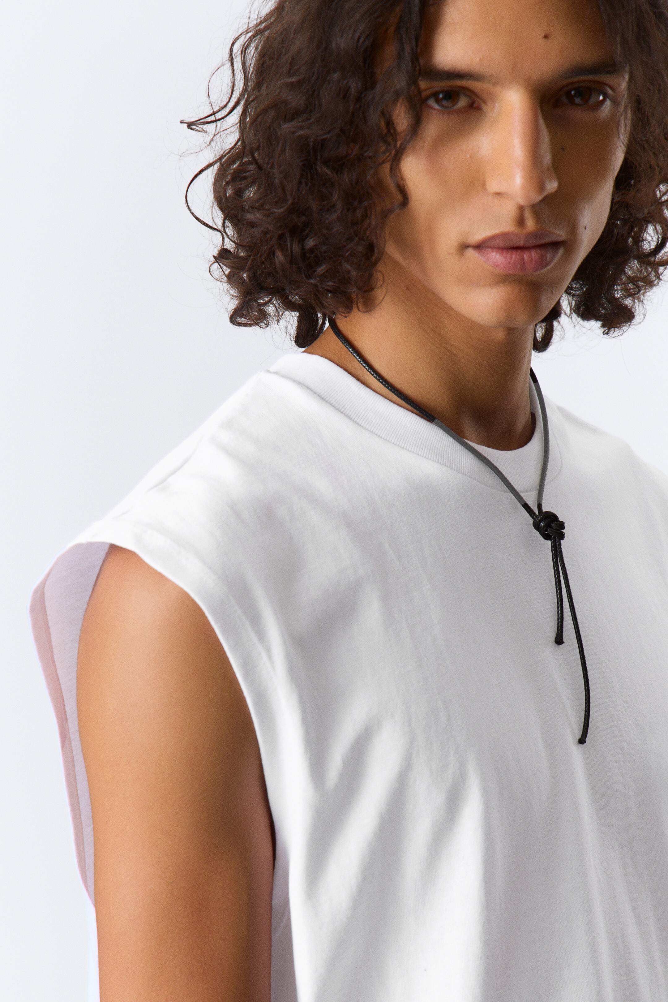 Grotere afbeelding bekijken: Een wit mouwloos topje wordt gedragen, iets schuin naar rechts gericht en vertoont een ronde hals en brede, strak gesneden armsgaten. Een zwart koordketting met een knoopdetail rust op de voorkant van het topje.