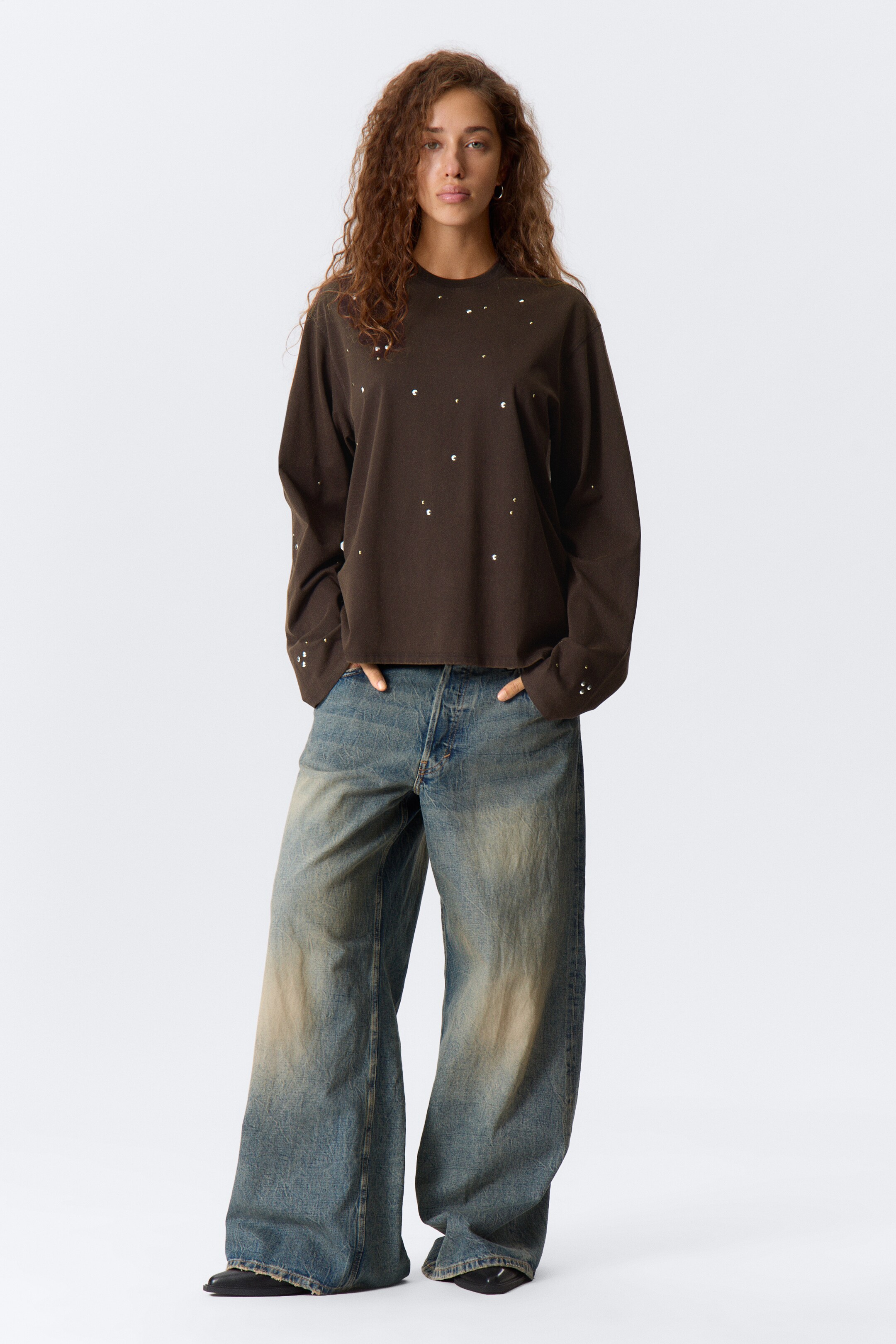 Visa större bild: En löst sittande, mörkbrun långärmad topp med en rund hals och spridda vita prickar bärs över vida blå jeans med omfattande blekning, presenterad från en framsida vy.