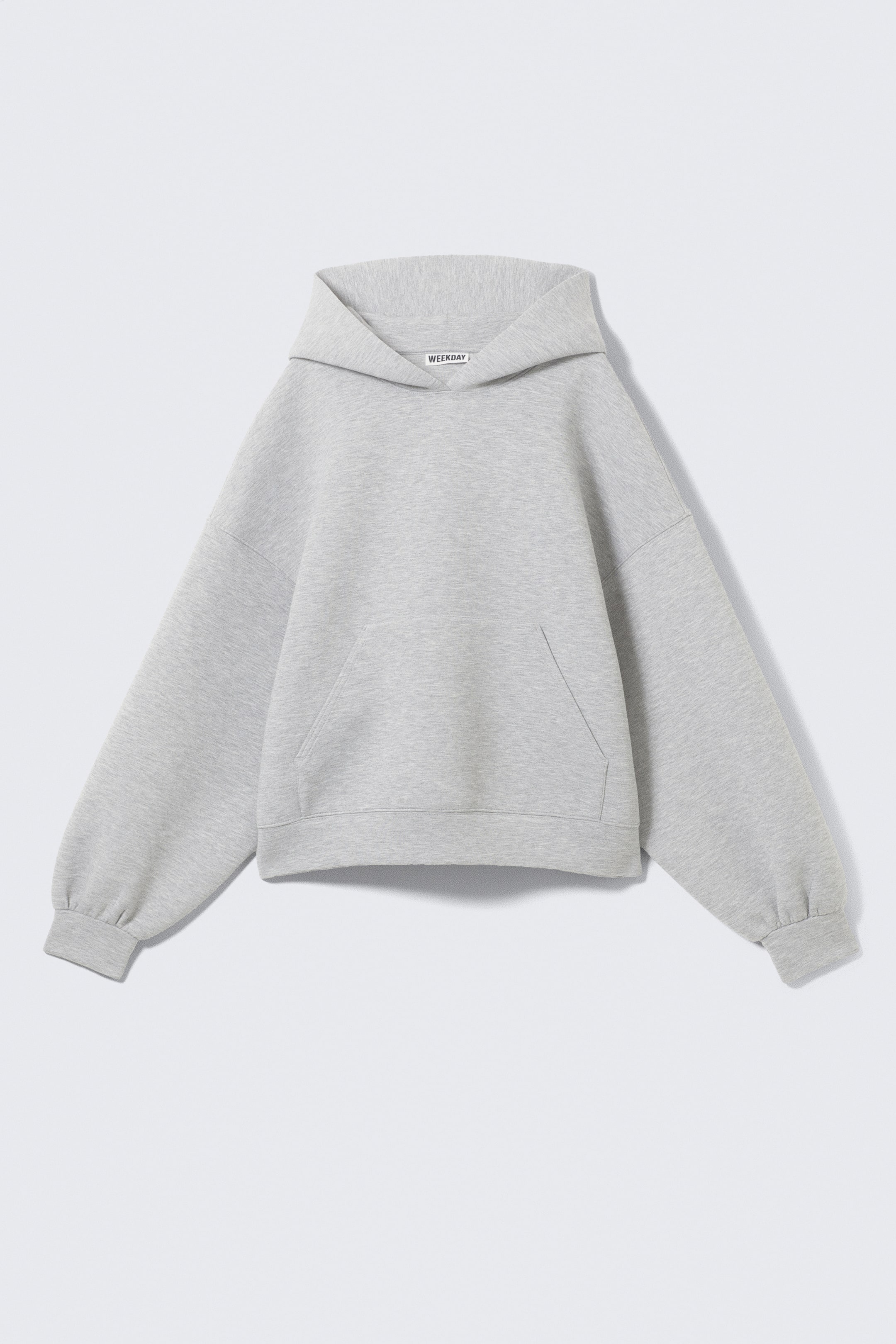 Visa större bild: En ljusgrå hoodie är vänd framåt, med en bred kragliknande huva och en synlig 'WEEKDAY'-märkesetikett. Den har överdimensionerade ärmar med samlade ribbade muddar, en stor känguruficka framtill och en ribbad fåll.