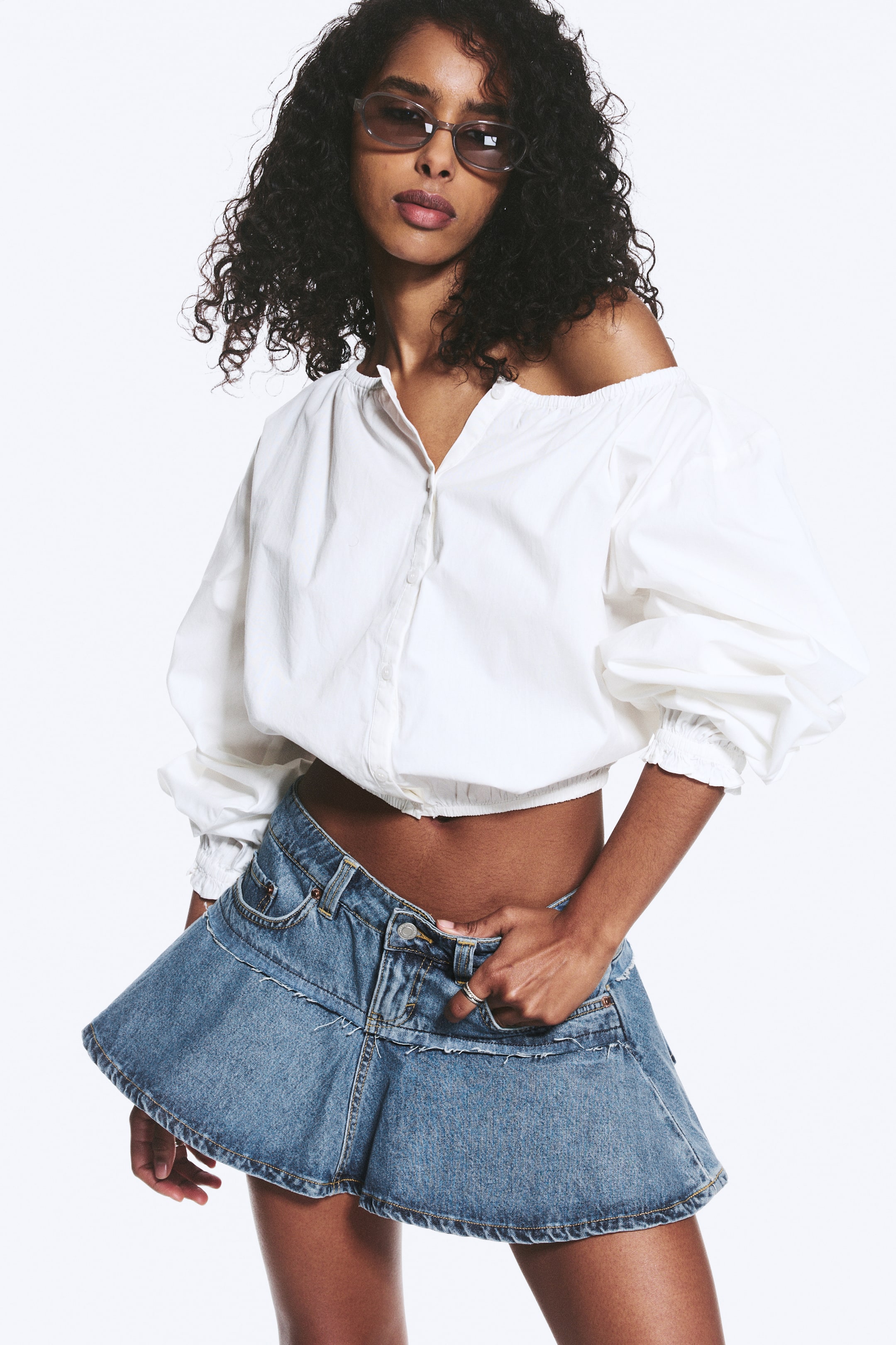 Grotere afbeelding bekijken: Een witte off-the-shoulder top met een bandkraag, een gedeeltelijk geopende knoopsluiting aan de voorkant, volumineuze lange mouwen met elastische manchetten en een elastische, korte zoom. Een blauwe denim low-rise minirok met een uitlopende pasvorm, zichtbare naden en een knoop- en ritssluiting aan de voorkant.