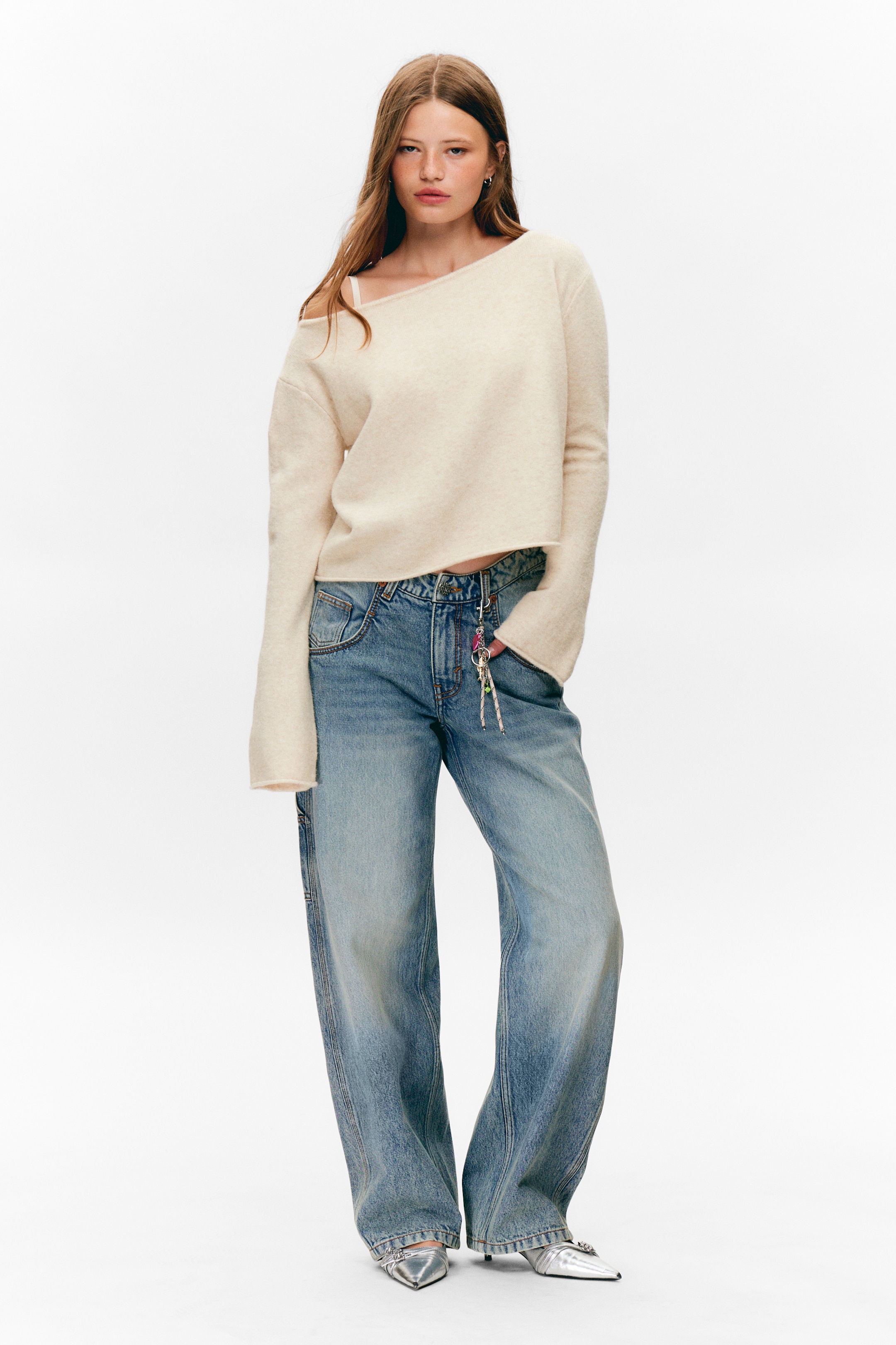 Ver imagem maior: Um camisola de cor creme, com textura suave, está a ser apresentada, apresentando um decote assimétrico fora do ombro no lado esquerdo do utilizador, mangas largas e uma bainha ligeiramente cortada e sem costura, revelando uma alça de sutiã branca visível. É combinada com jeans de perna larga e lavagem clara que têm uma aparência desbotada e um porta-chaves colorido pendurado num passador de cinto, juntamente com mules de salto prateado com bico pontiagudo.