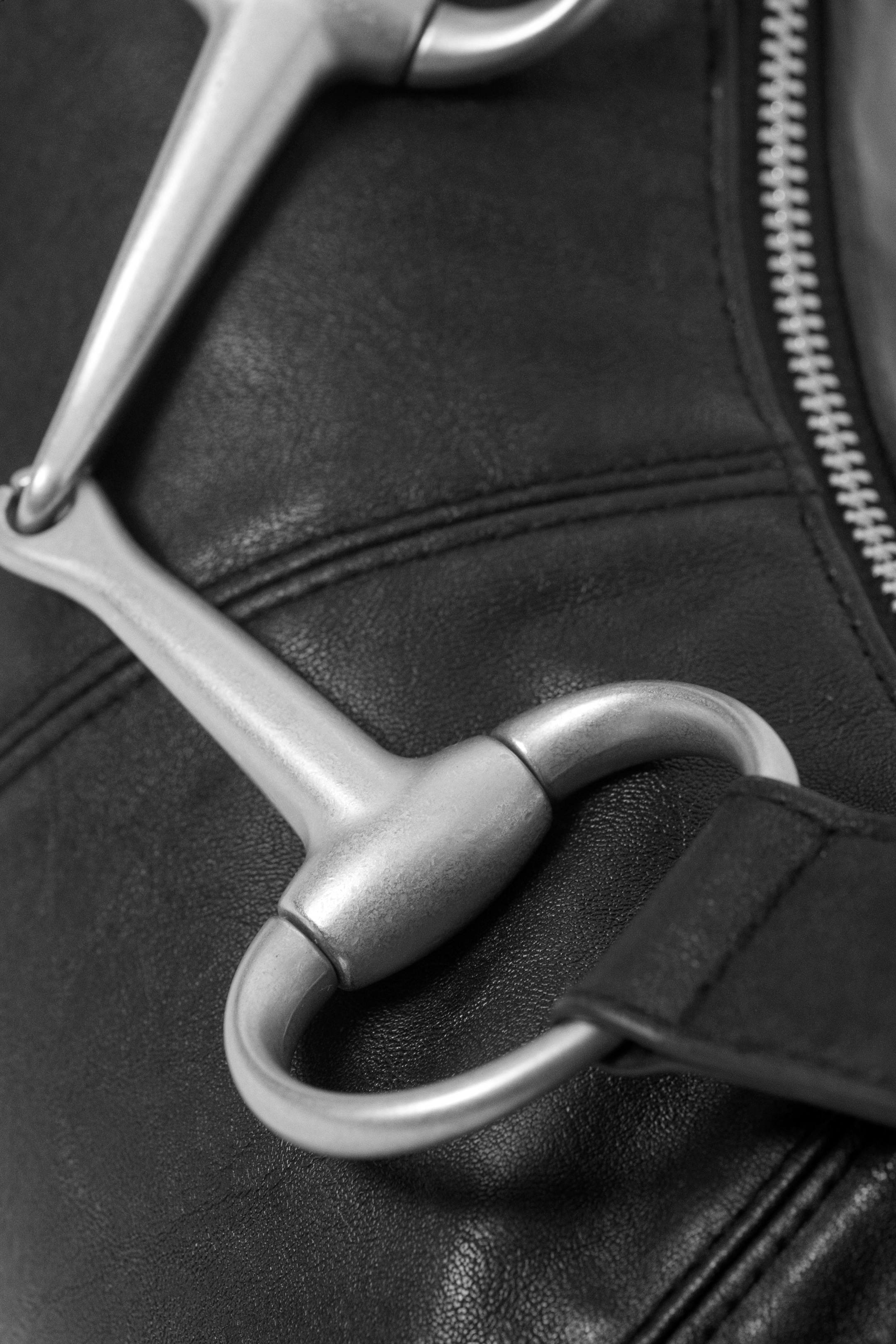 Visa större bild: En hästbett i silvertonad metall, med ett ledat munstycke och en borstad finish, är fäst vid en svart, texturerad läderyta som också visar en silvertonad dragkedja med framträdande tänder.
