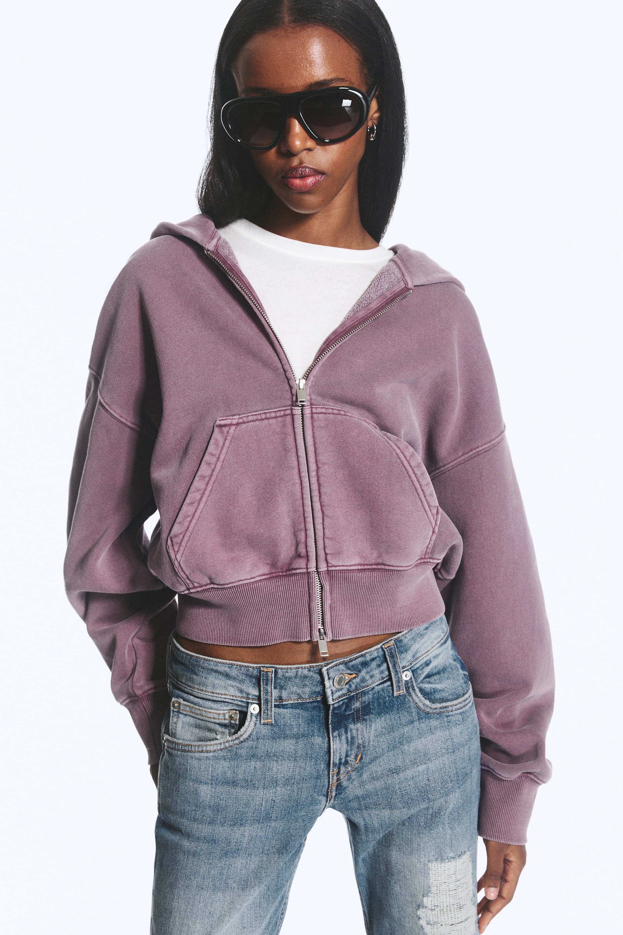 Agrandir l'image: Un hoodie court à capuche zippé de couleur violet délavé est présenté de face, avec une fermeture éclair argentée sur toute la longueur, une poche kangourou, une capuche, des poignets côtelés et un ourlet côtelé, superposé sur un t-shirt à col rond blanc.