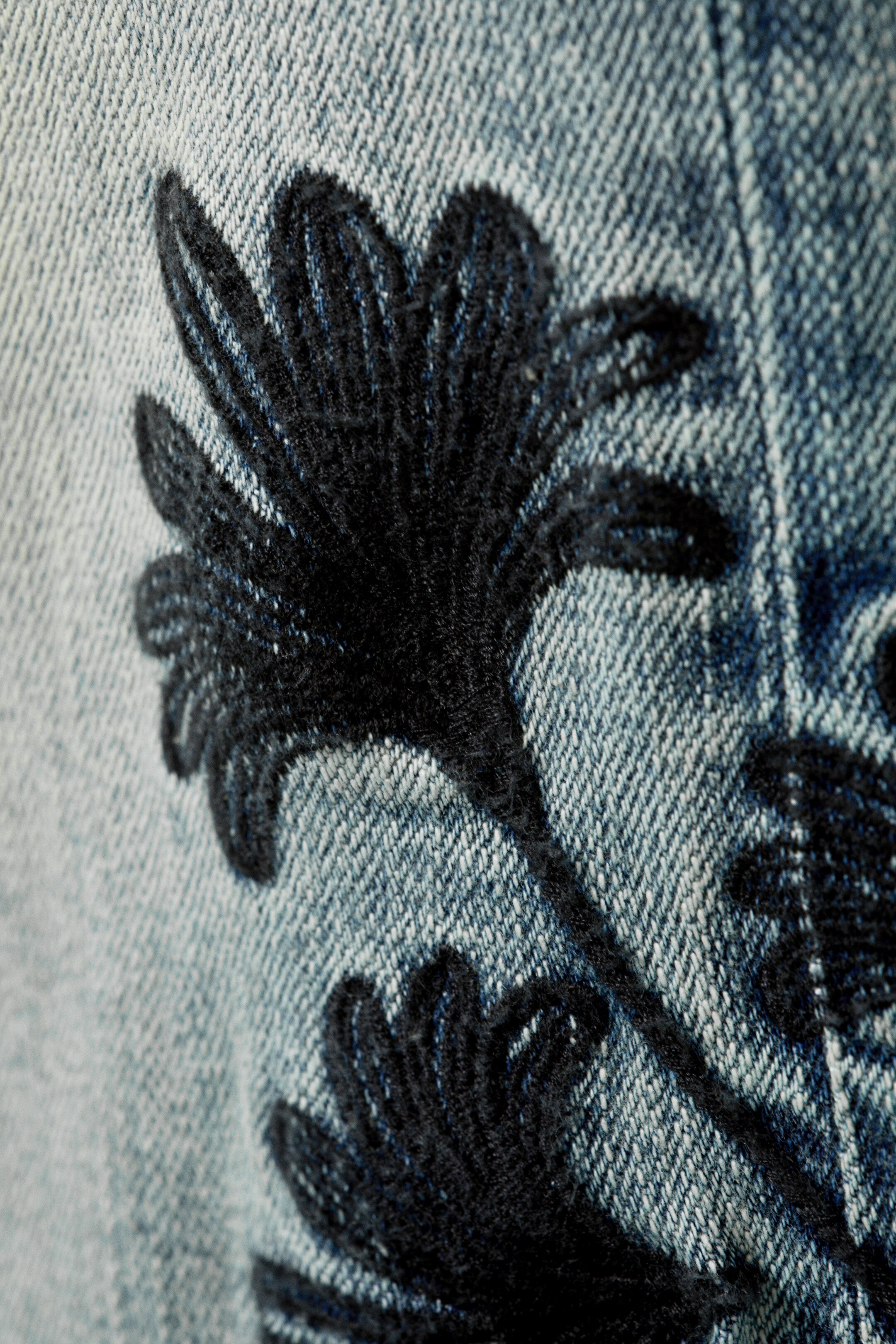 Größeres Bild ansehen: Ein hellblauer Denimstoff wird in einer Nahaufnahme gezeigt, mit schwarzer, erhabener Stickerei. Die Stickerei bildet ein dichtes, strukturiertes Blumen- oder Blattmuster über den Stoff.
