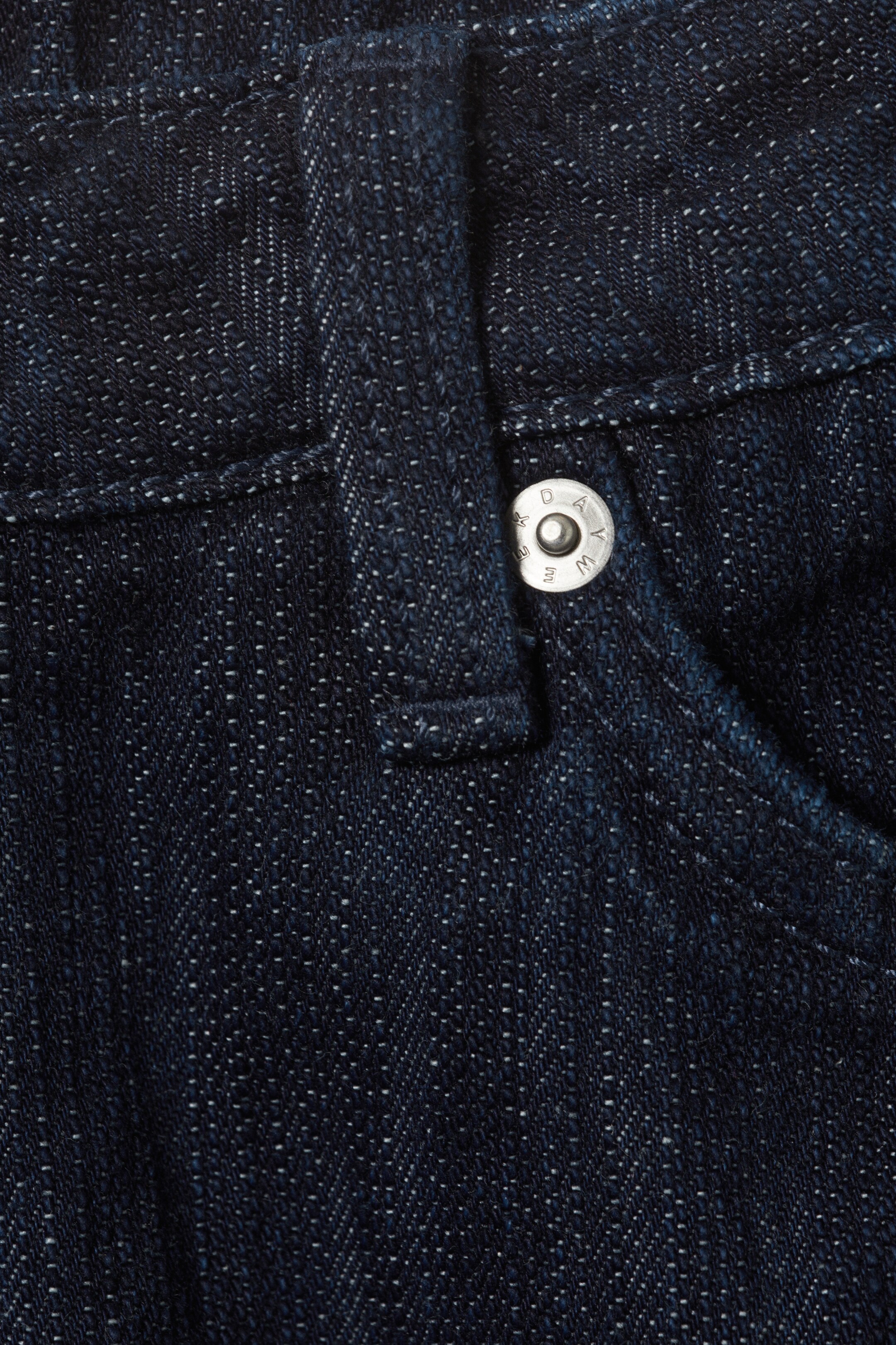 View larger image: Dark blue denim fabric is displaying a silver-colored metal button on the front. A belt loop is positioned to the left of the button, and a pocket opening is visible to its right, with the fabric showing a textured weave and lighter flecks.