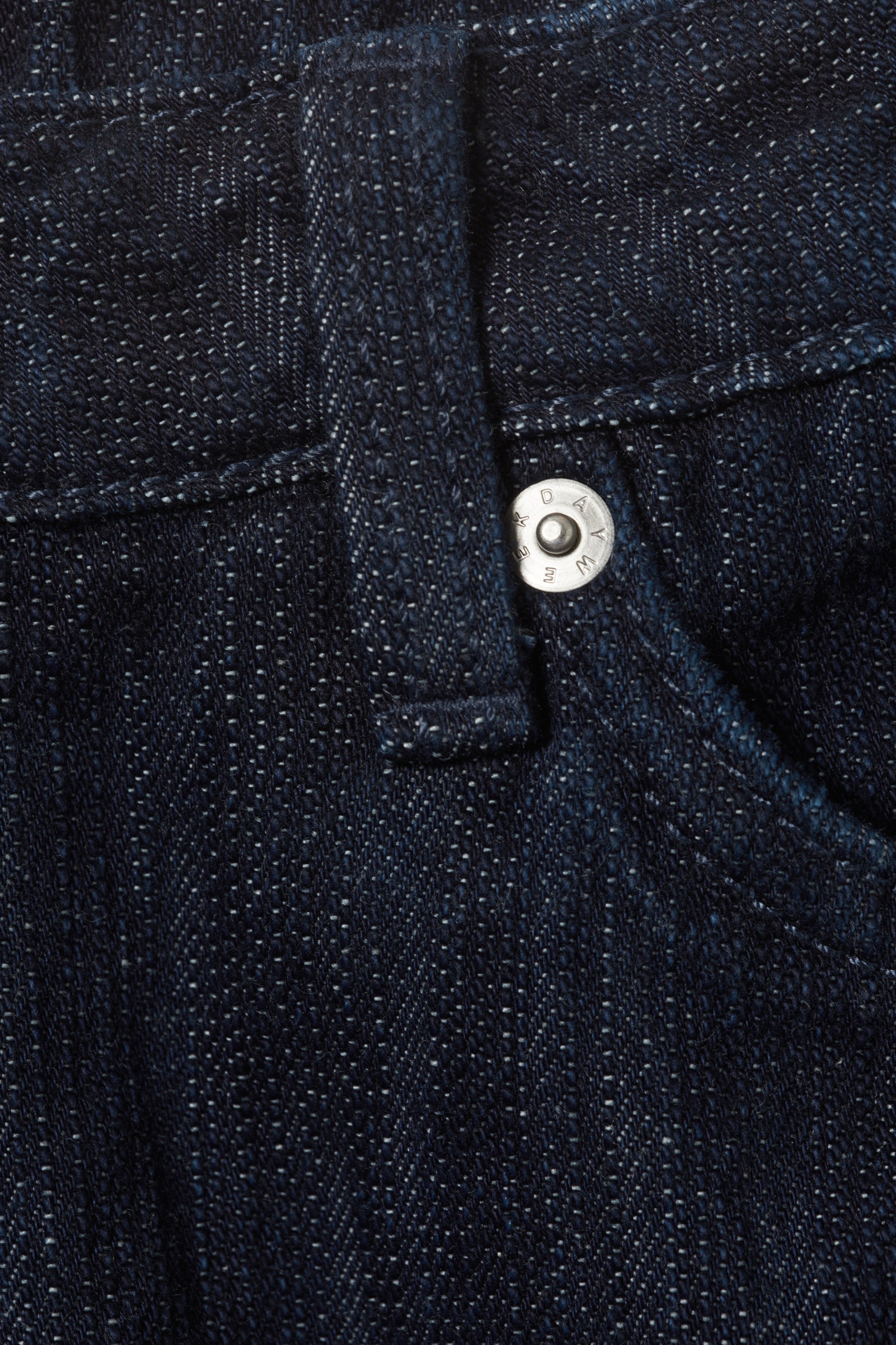 Grotere afbeelding bekijken: Donkerblauwe denimstof toont een zilverkleurige metalen knoop aan de voorkant. Een riemlus bevindt zich links van de knoop en een zakopening is zichtbaar aan de rechterkant, waarbij de stof een getextureerde weving en lichtere vlekjes vertoont.