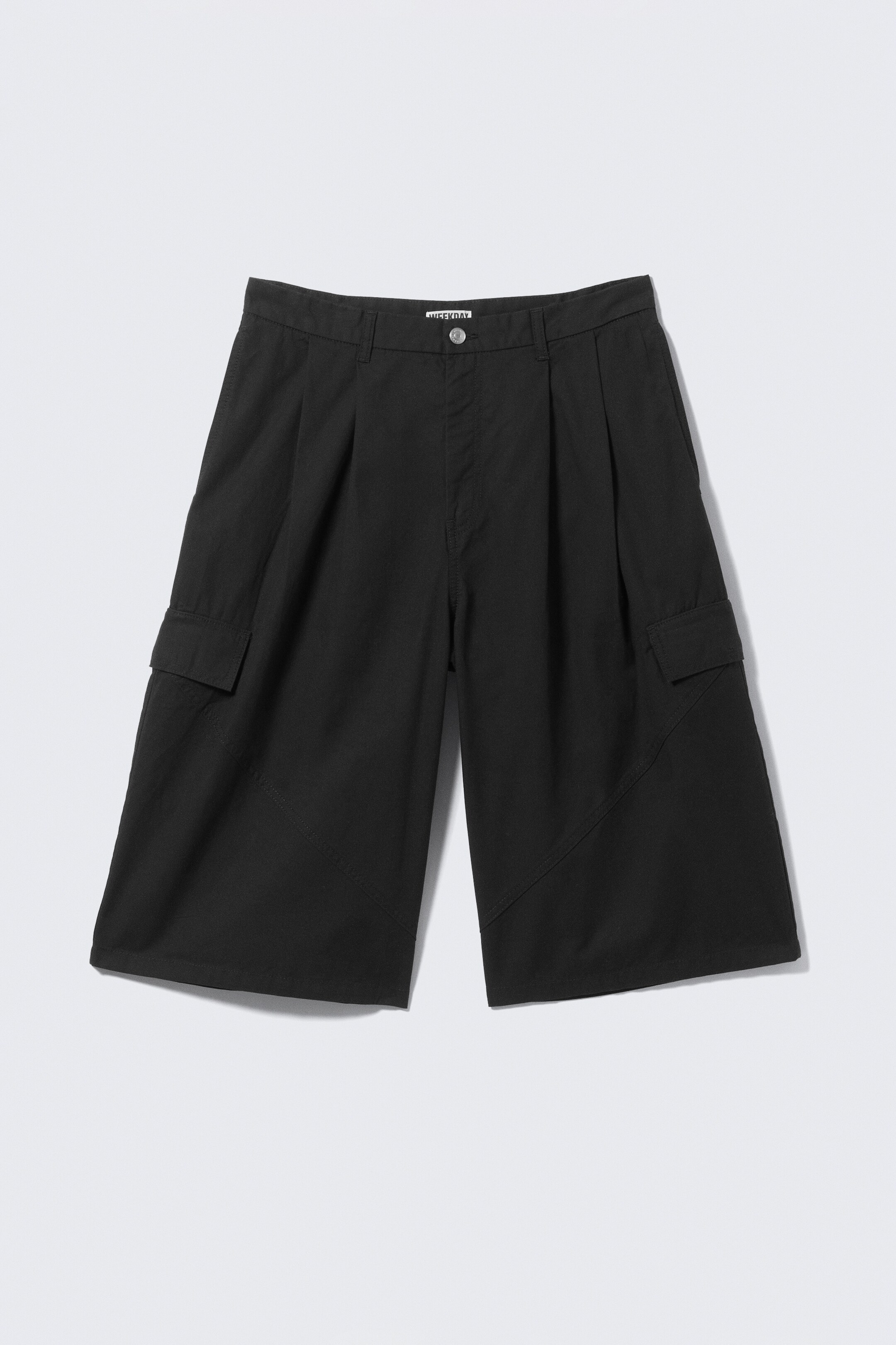 Ingrandisci l'immagine: Un paio di pantaloncini neri, taglio ampio, che arrivano al ginocchio sono presentati dal FRONTE. Realizzati in twill di cotone, i pantaloncini presentano pieghe sul davanti, cuciture laterali angolate e tasche con pattina sulle cosce. Passanti per cintura e una chiusura con zip e bottone completano la vita strutturata.